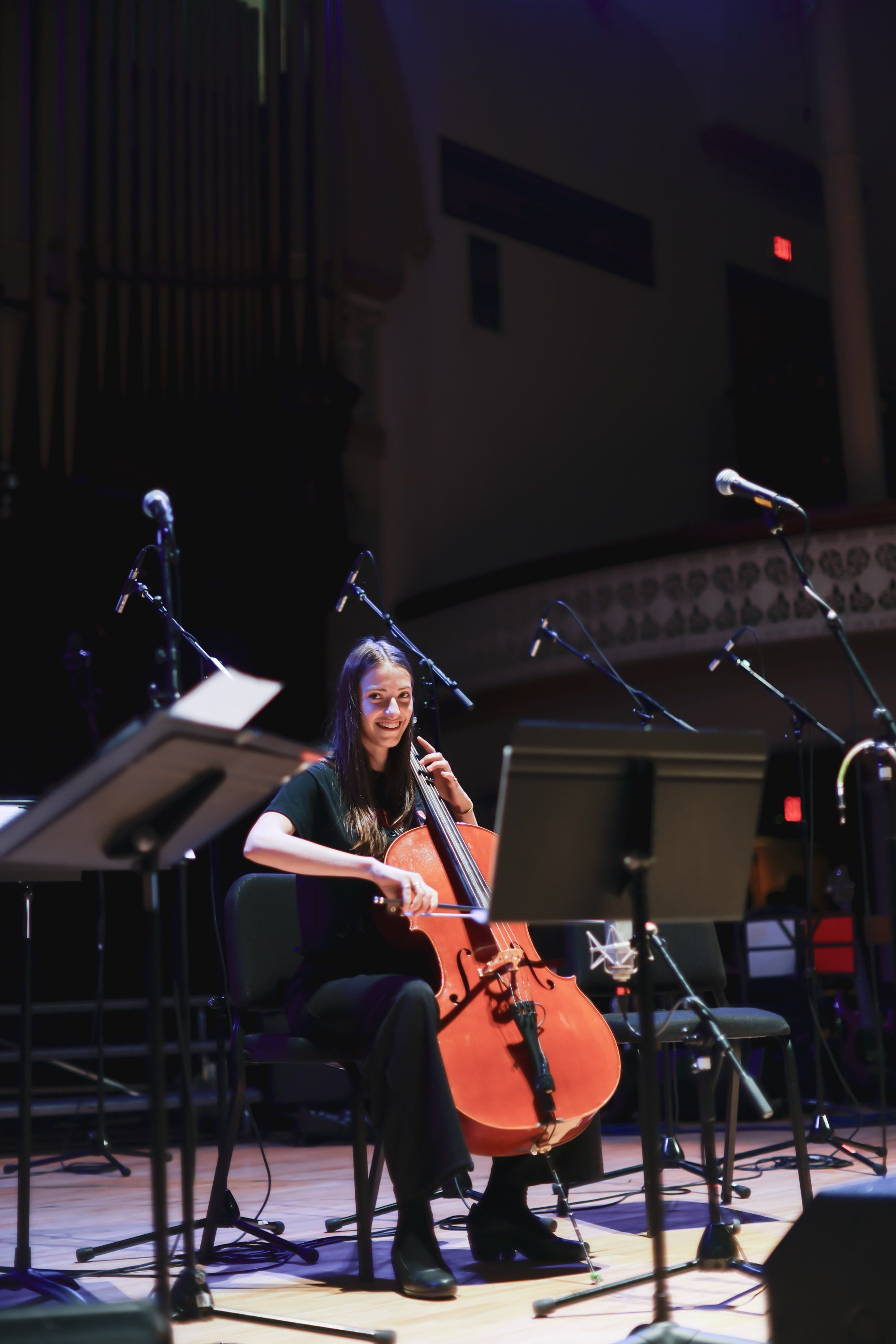 Cellist performing solo on stage under warm spotlight, with music stands and microphones visible in the dark concert hall