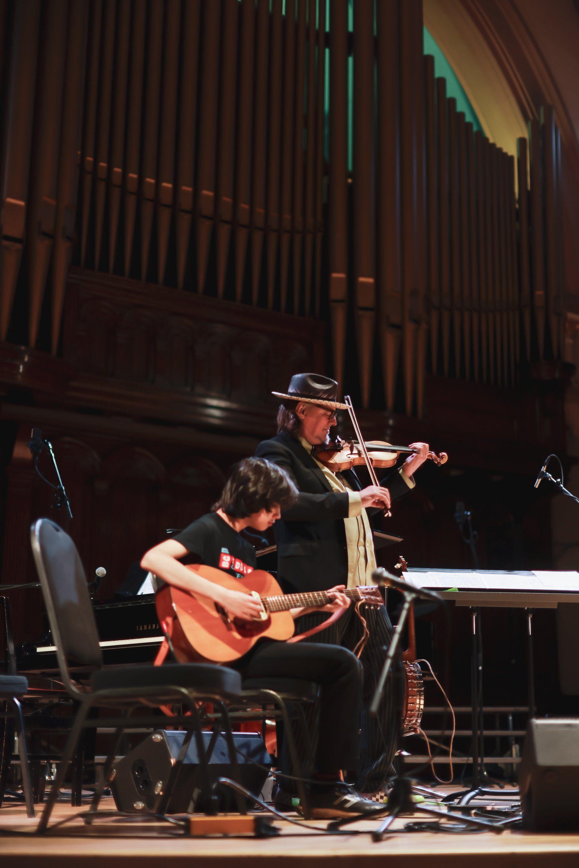 Two musicians perform on stage in warm lighting: one playing violin wearing a hat, the other on acoustic guitar, with a grand organ visible in the background