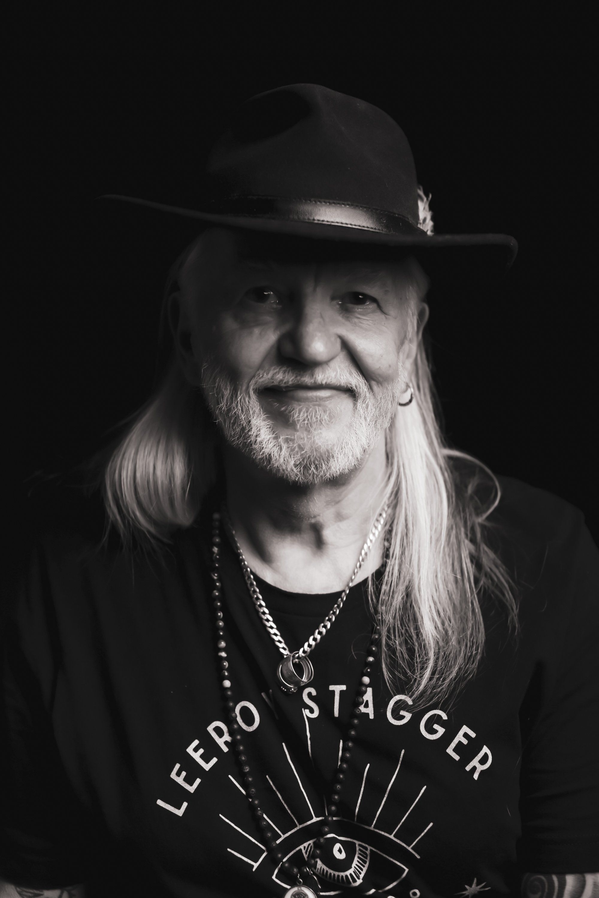 Long-haired man in cowboy hat and graphic tee smiles at camera against dark background, dramatic side lighting