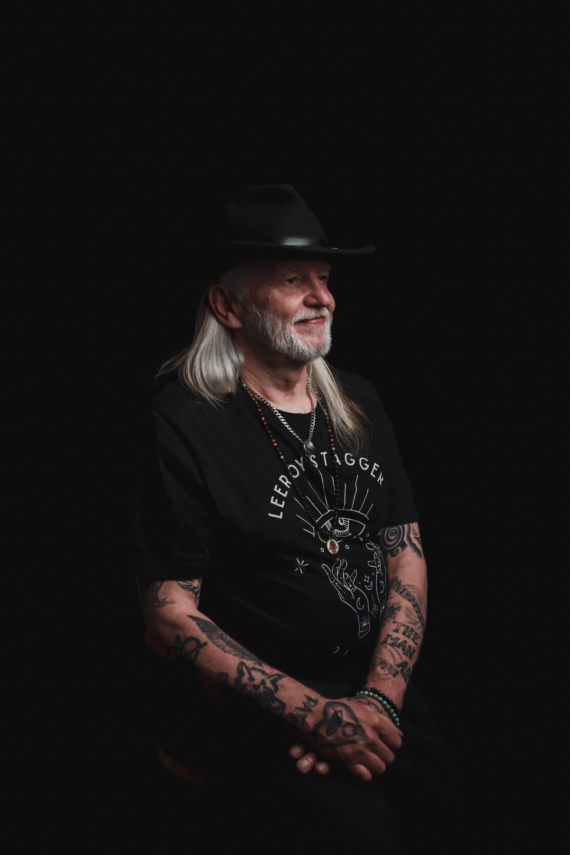 Long-haired man in black cowboy hat and graphic tee with arm tattoos, posed against dark background with dramatic side lighting