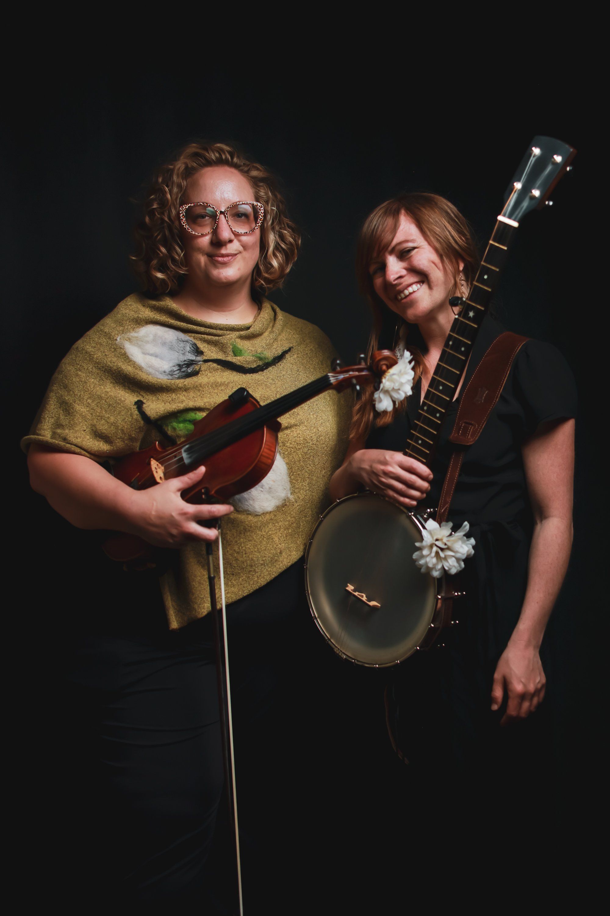 Two musicians smile warmly against a dark background, one holding a mandolin in a yellow shirt, the other with a banjo wearing dark clothing