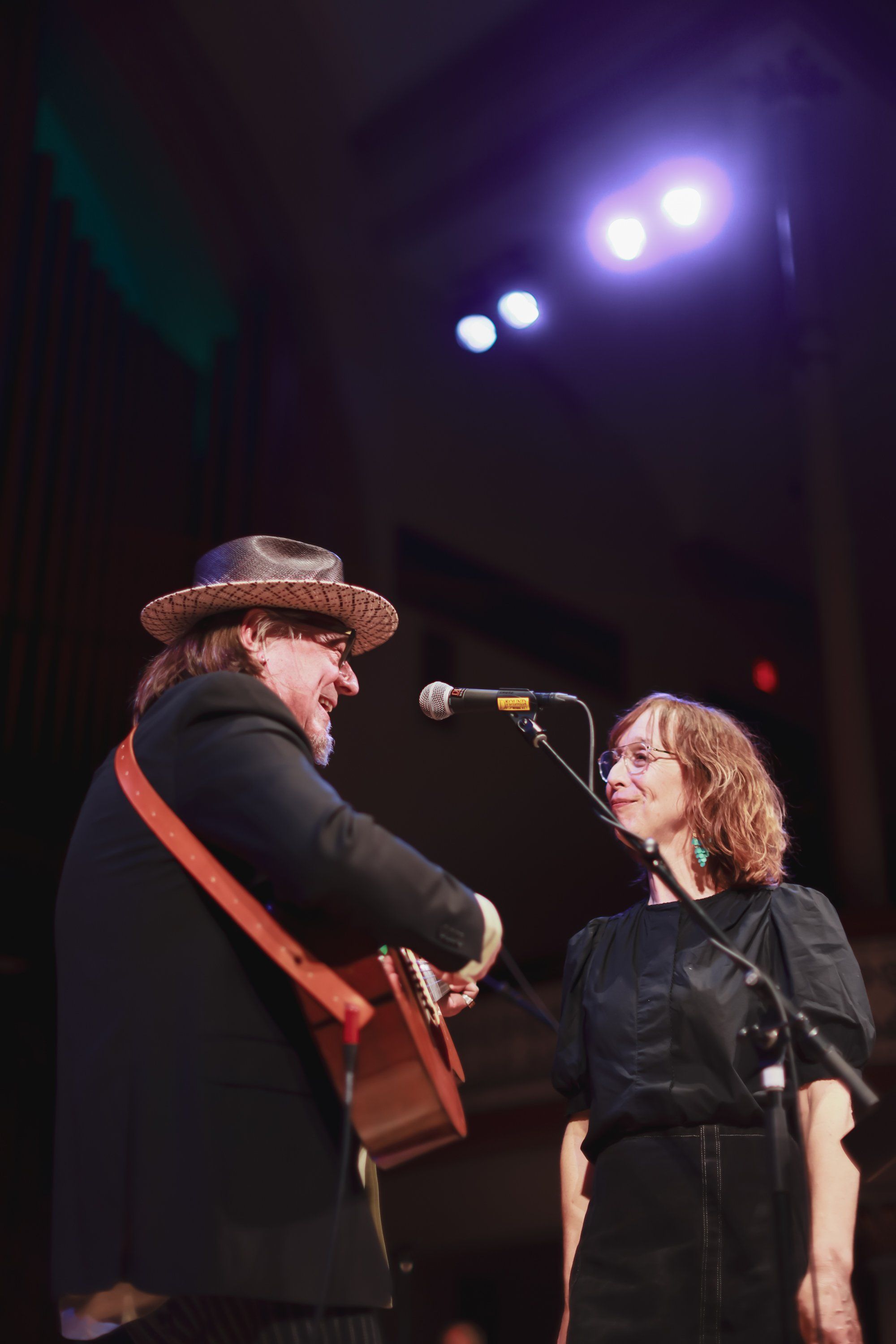 Two performers share a microphone on a dimly lit stage, one wearing a brown hat and rust-colored scarf, the other in black, with dramatic overhead lighting a…