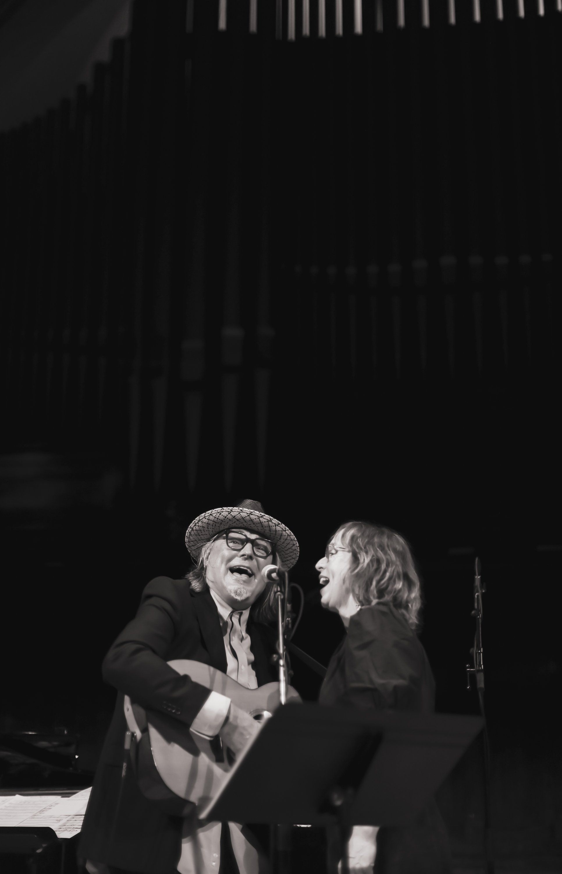 Two musicians sharing a moment on stage, one wearing a hat and glasses playing guitar, the other in profile, captured in warm spotlight against dark surround…