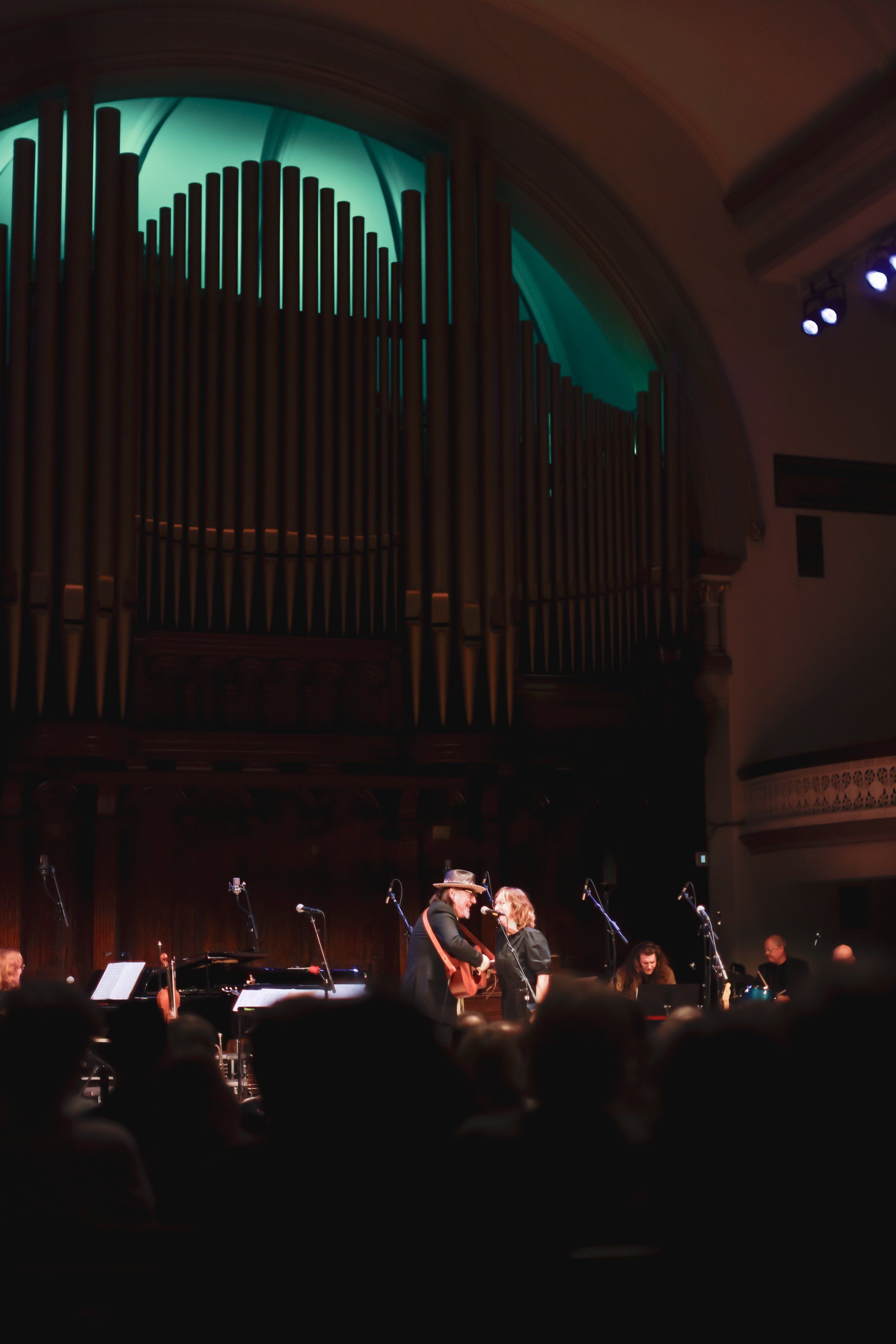 Musicians perform on stage beneath a glowing green pipe organ in a darkened concert hall with warm spotlights