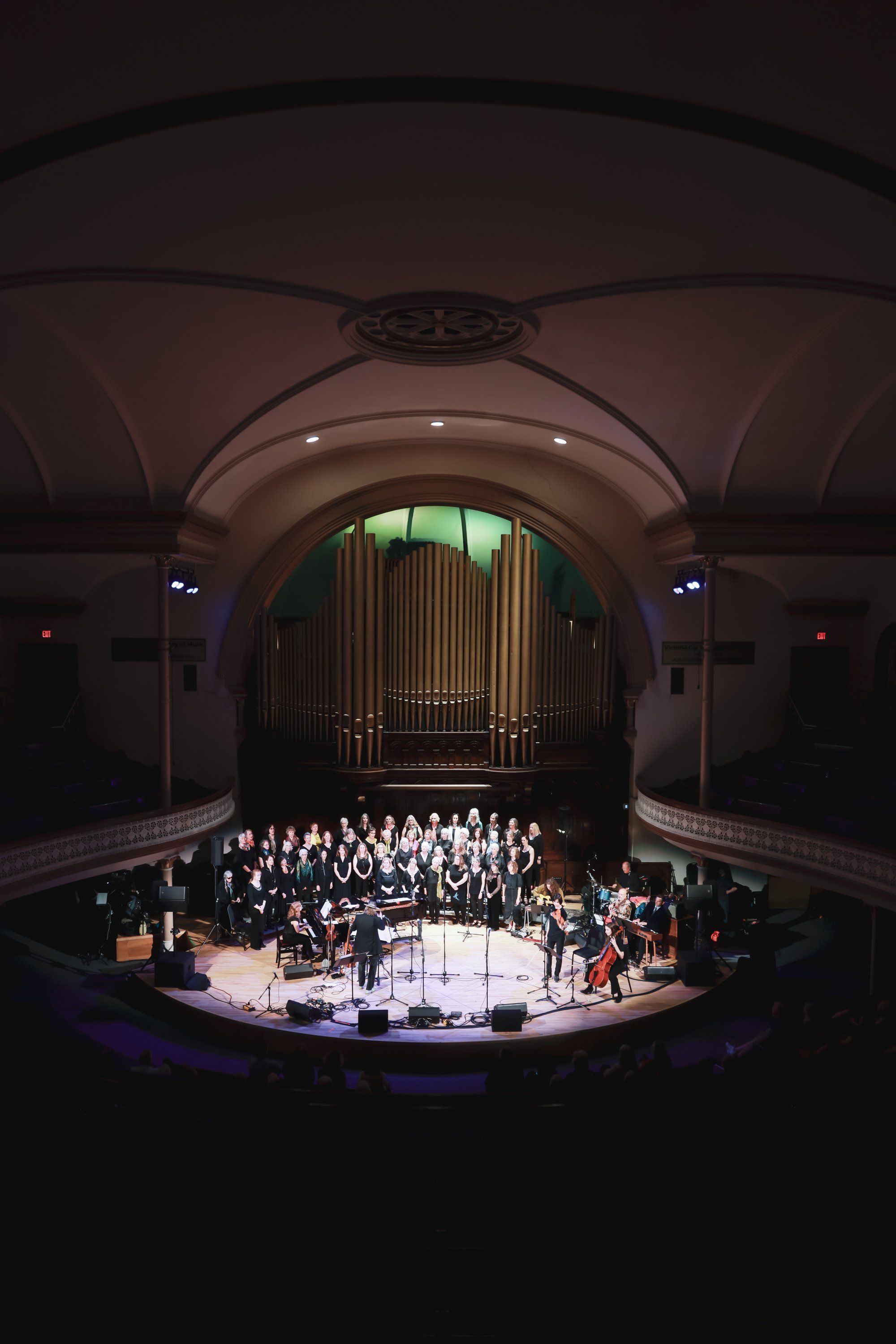 Orchestra and choir perform on an illuminated stage beneath a grand organ and arched ceiling in an elegant concert hall