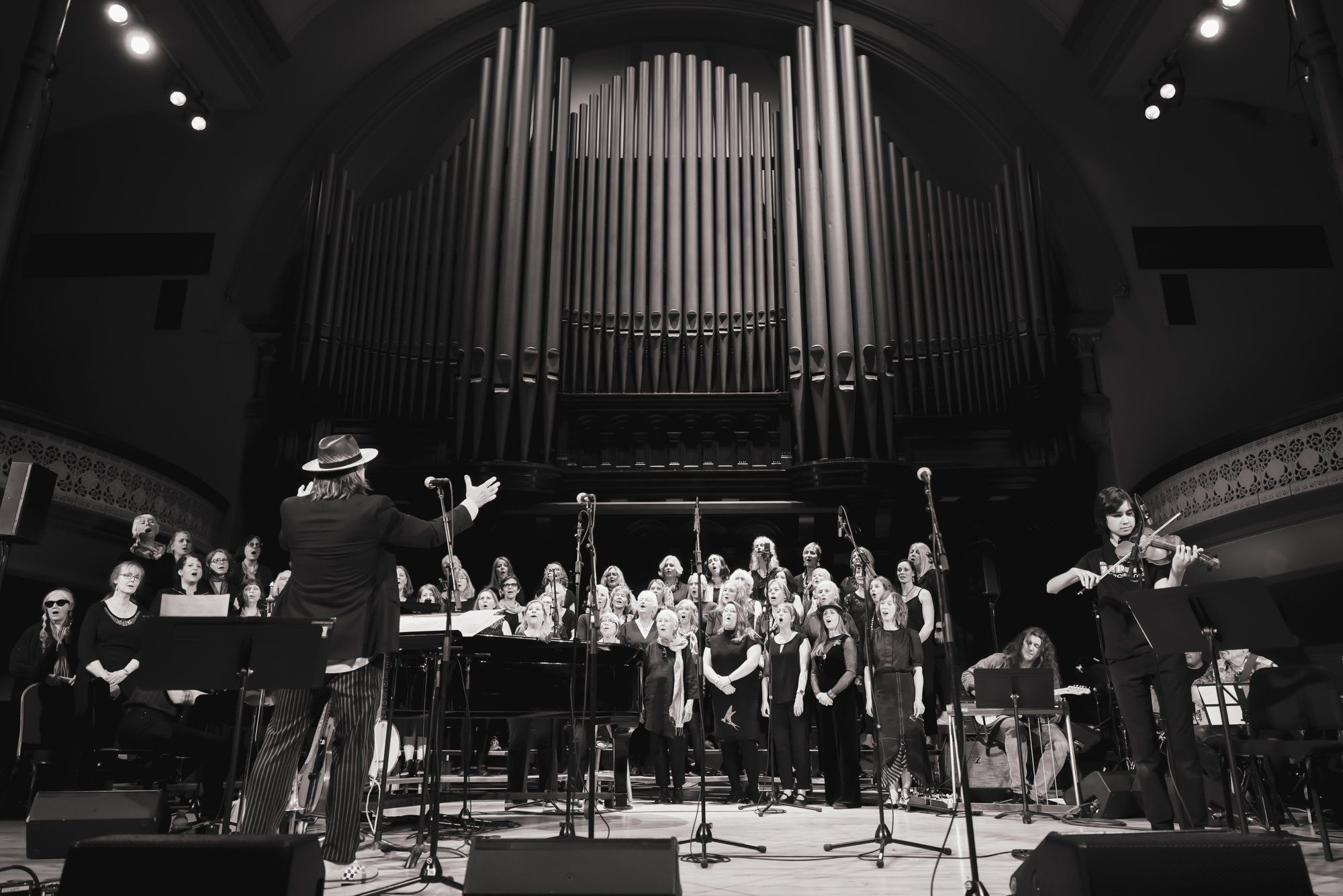 Conductor leading a large choir and orchestra on stage beneath a towering pipe organ in black and white
