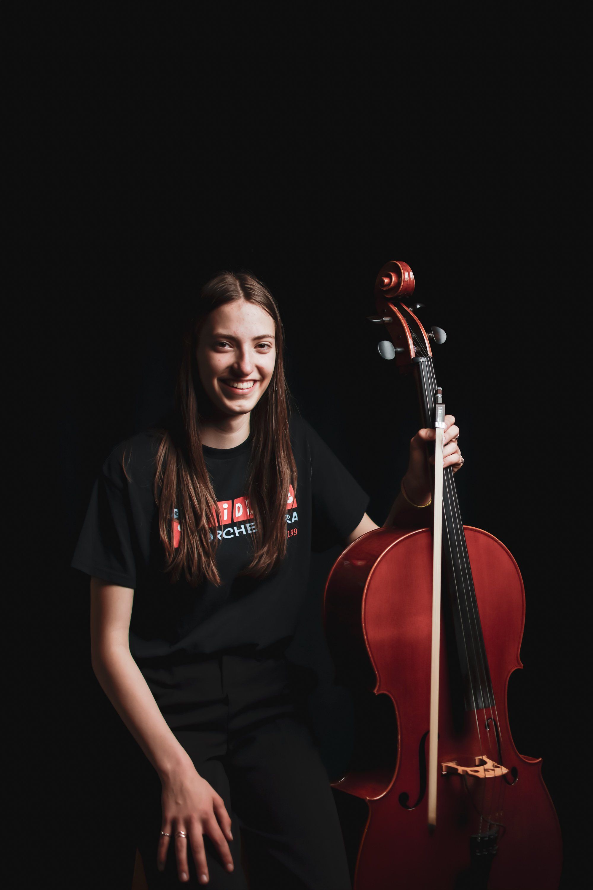 Young cellist with long dark hair smiles beside her instrument against a black background, warmly lit
