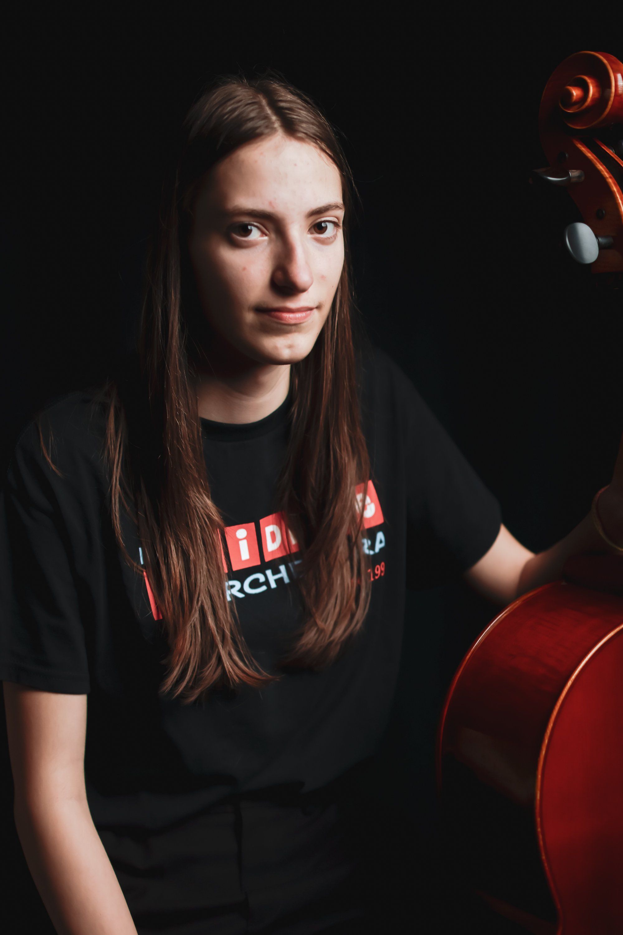 Young musician in black shirt smiles beside red cello against dark background, warm portrait lighting