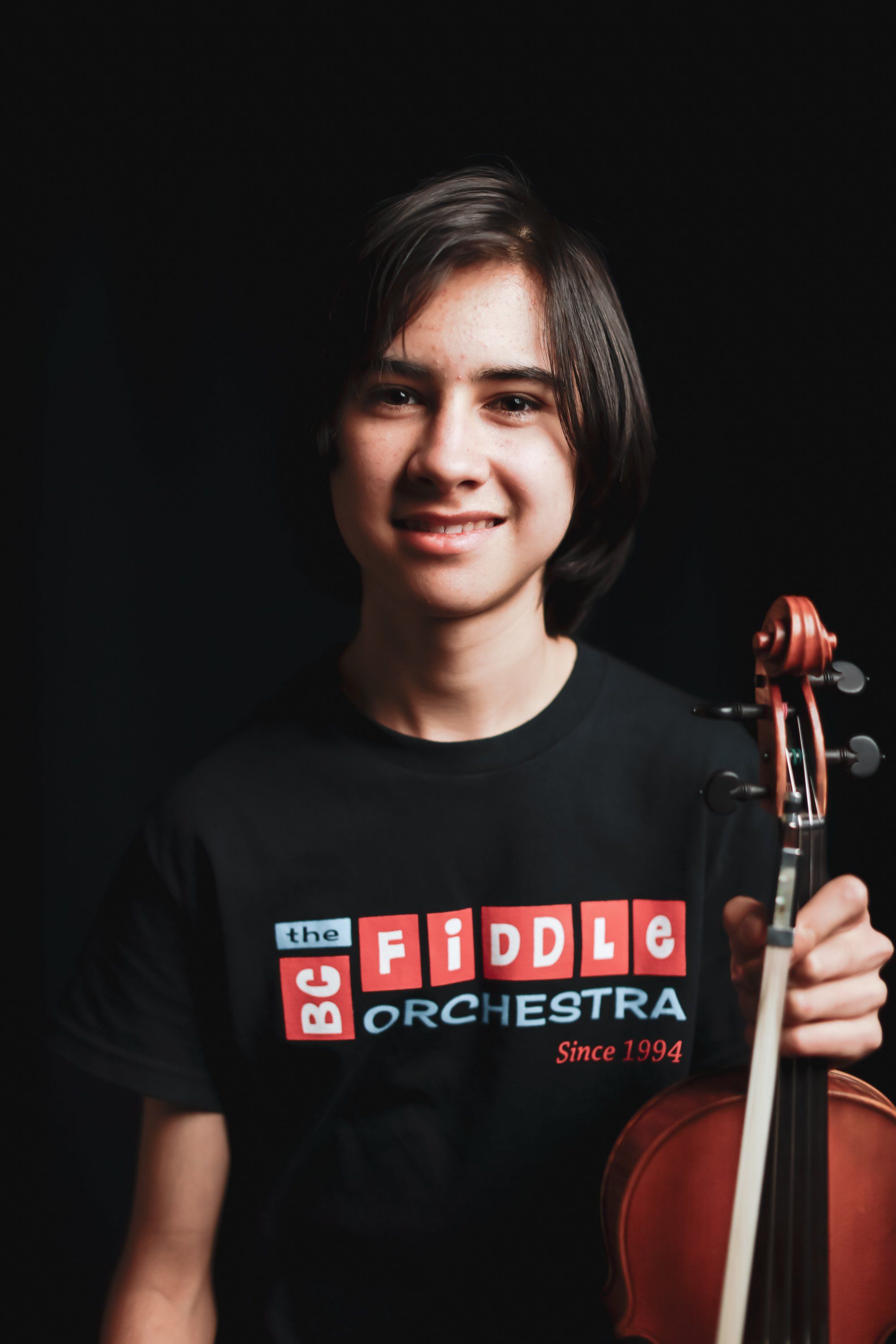 Young violinist holding instrument against black background, wearing Fiddle Orchestra t-shirt, smiling warmly at camera