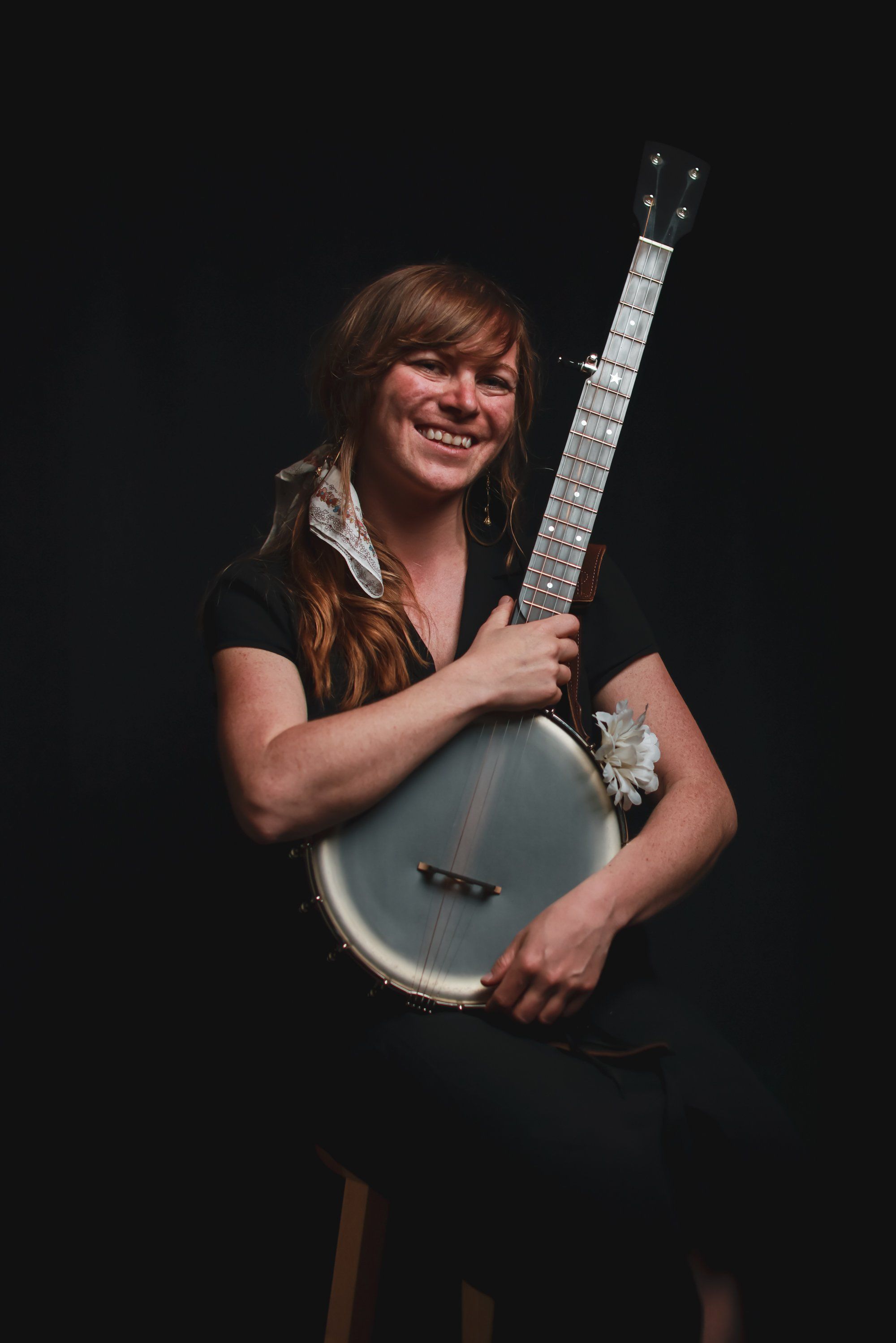 Smiling musician holds a white banjo against a black background, wearing a dark shirt with warm stage lighting