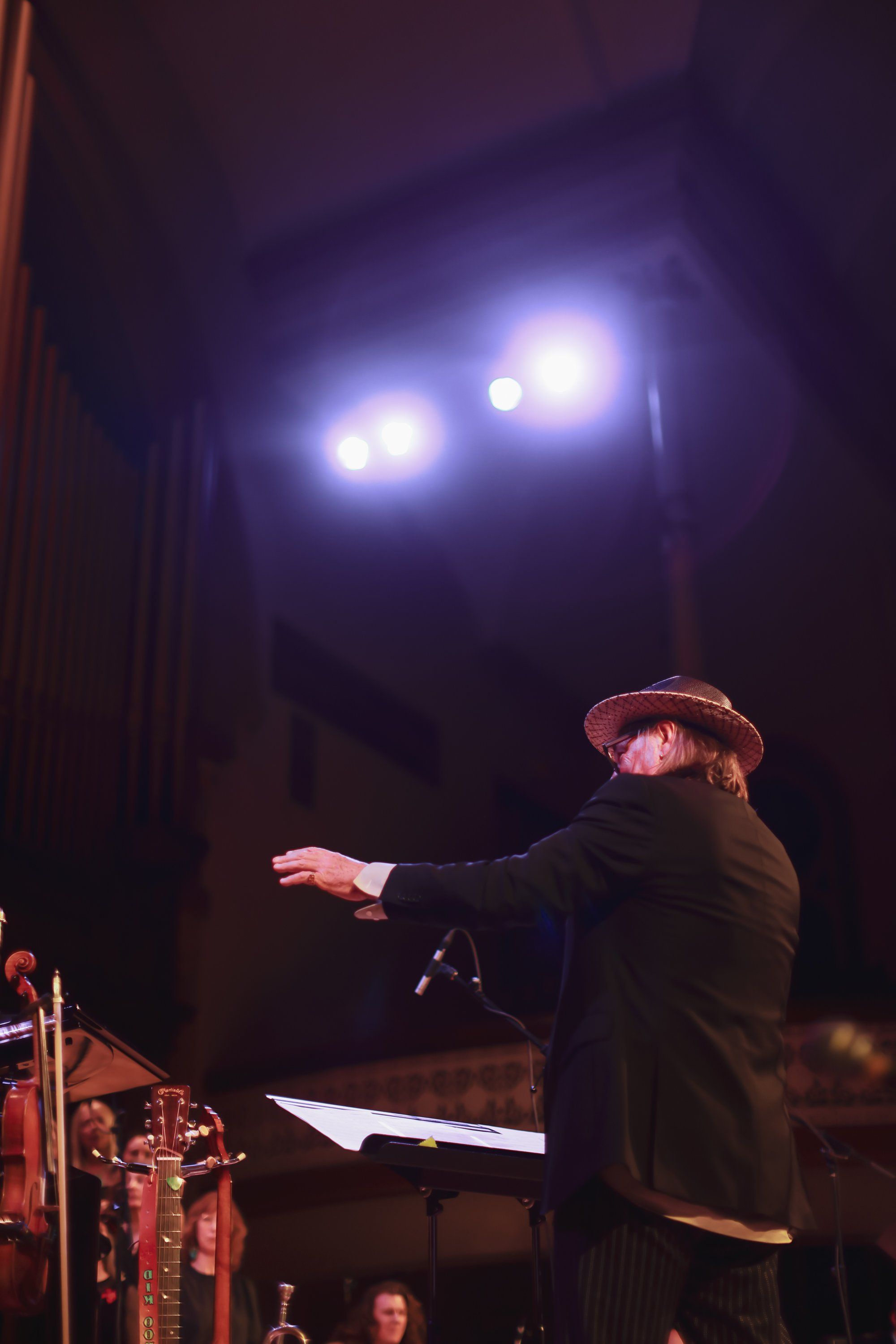 Conductor in dark coat gestures expressively on stage under bright spotlights, with orchestra drums visible below