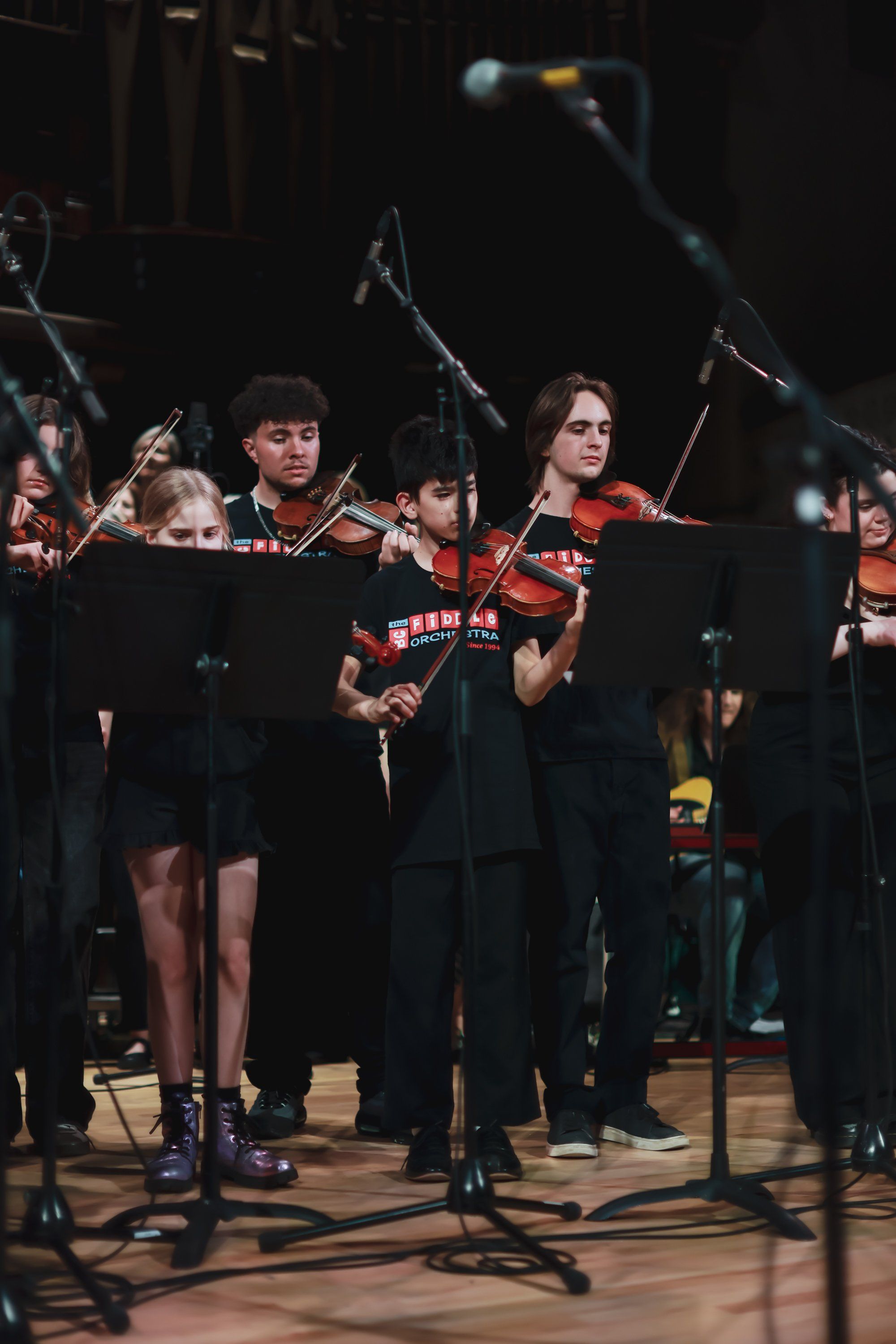 Orchestra musicians in red and black uniforms perform on stage under dramatic stage lighting during an evening concert