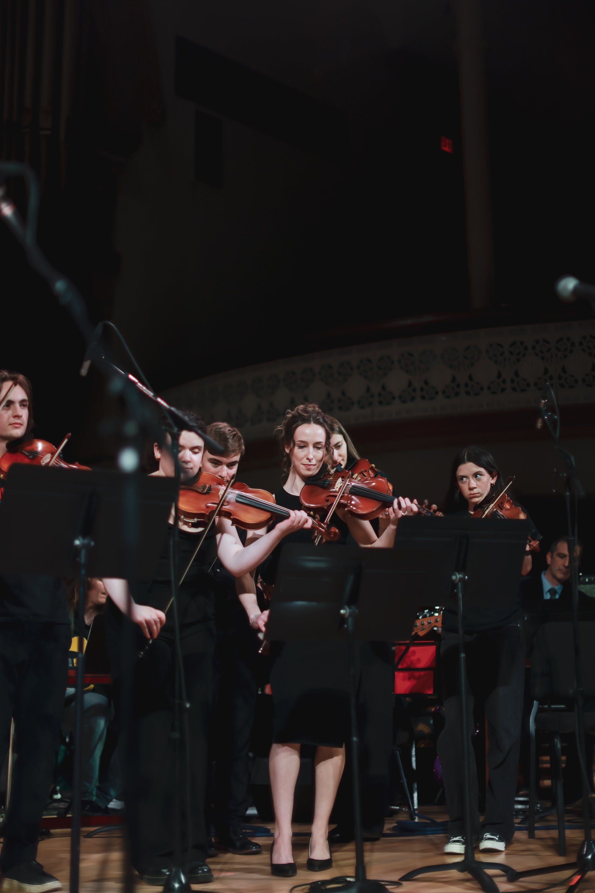 Young violinists in black concert attire perform on a stage lit by warm spotlights during an ensemble performance