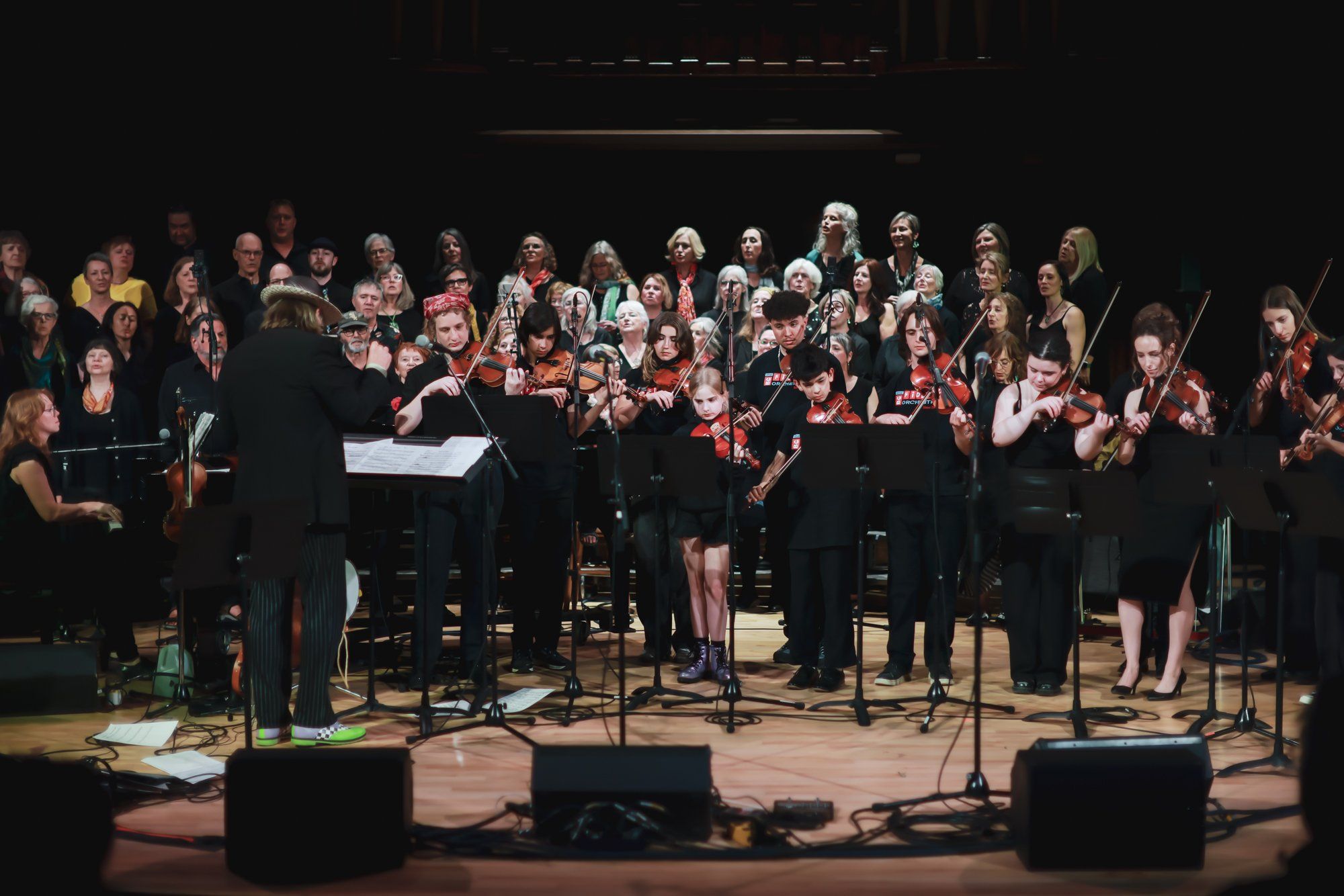 Large choir in formal black attire performs on stage under warm lights, conductor leading from podium, intimate concert setting