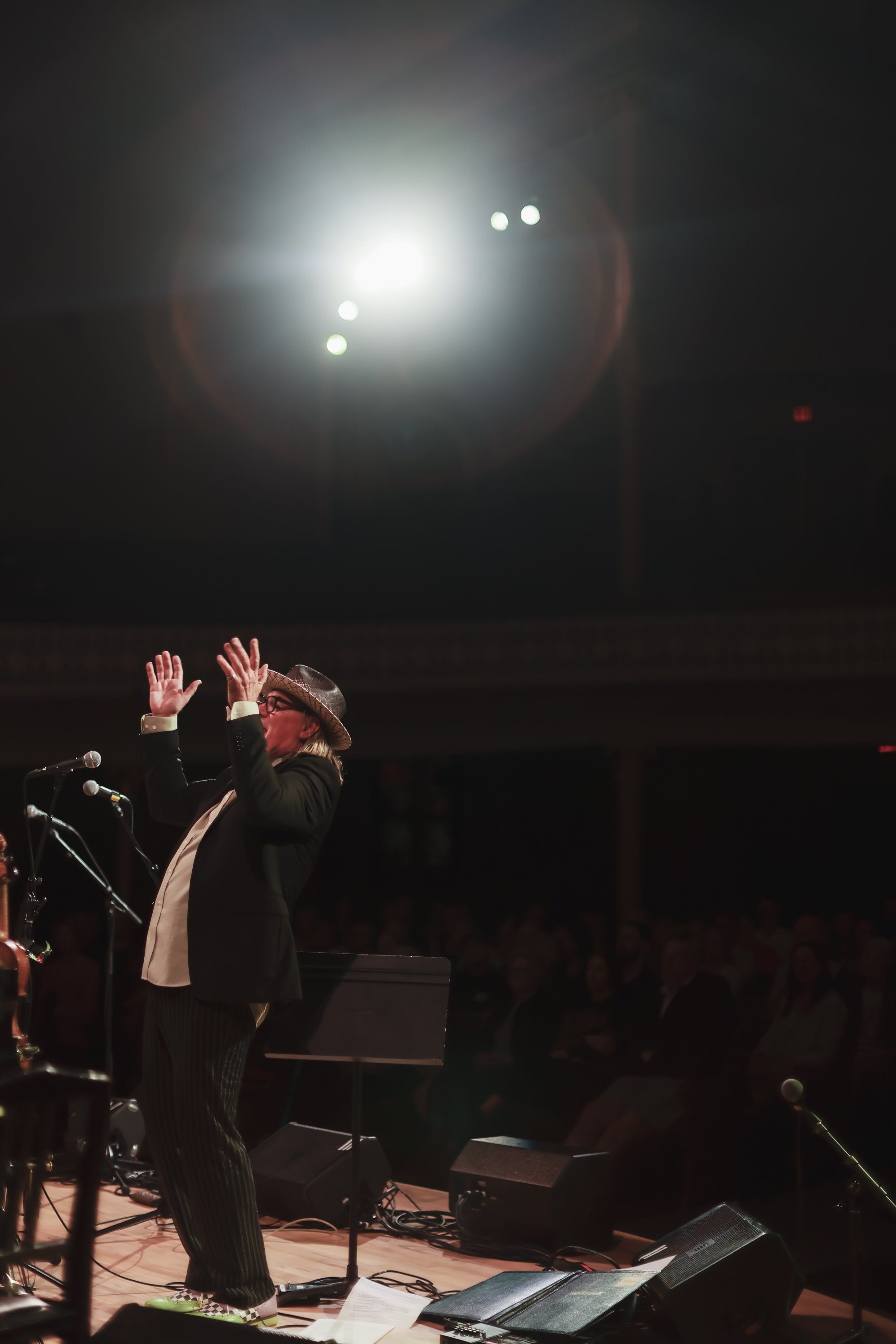Performer with hands raised on darkened stage beneath bright overhead lights, intimate concert atmosphere