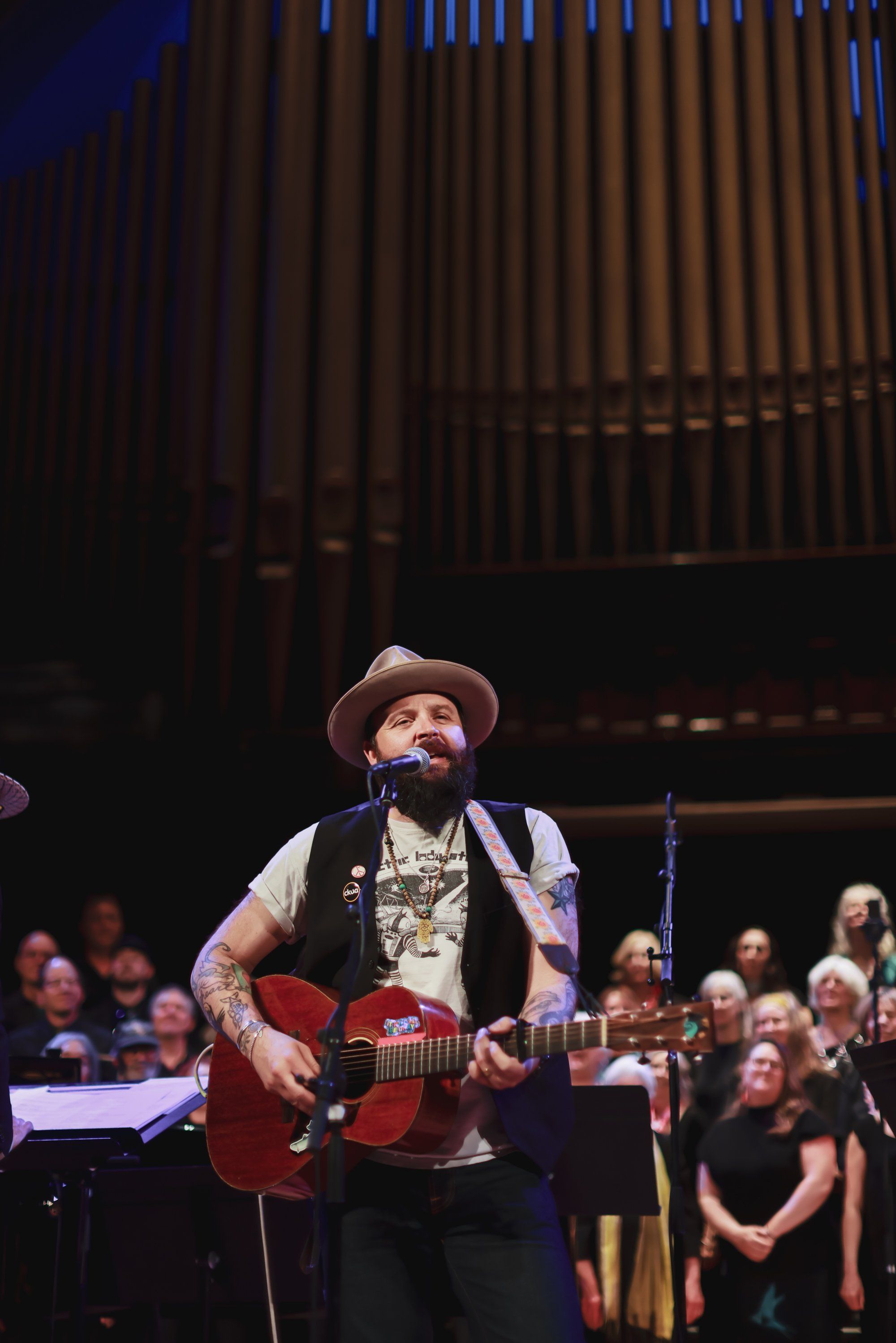 Bearded musician in cowboy hat and red guitar performs under warm stage lights with choir members in background