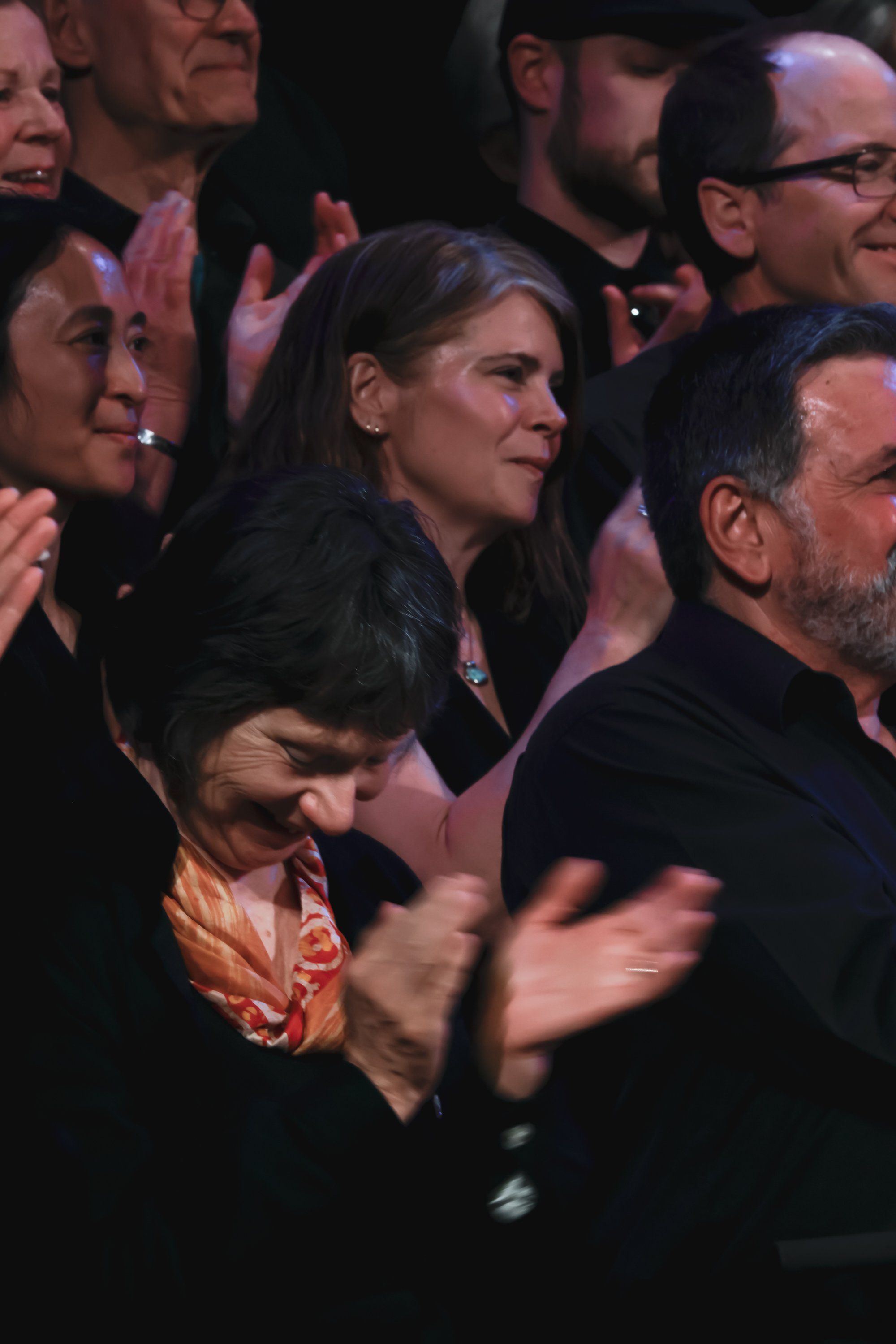 Audience members smile and applaud during an evening concert, bathed in warm stage lighting against a dark background