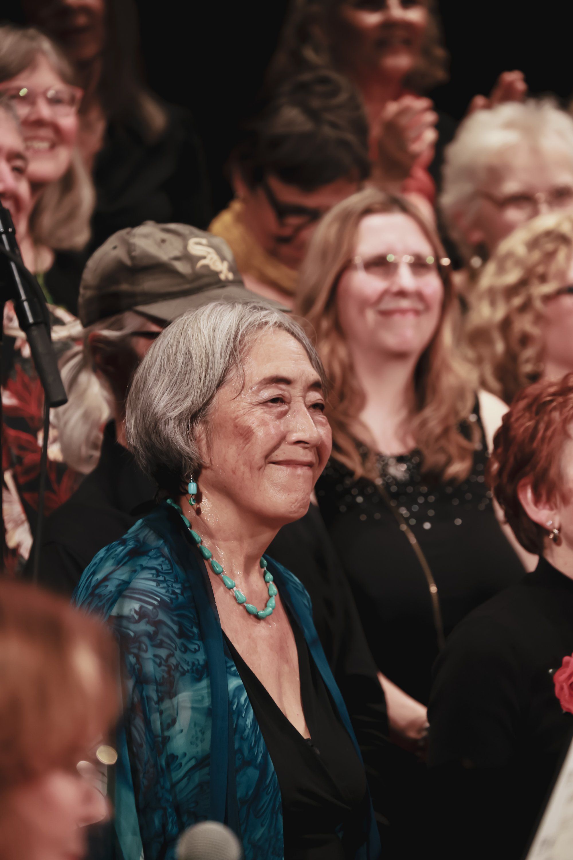 Gray-haired woman in teal jacket smiles warmly amid seated audience members at an indoor concert venue