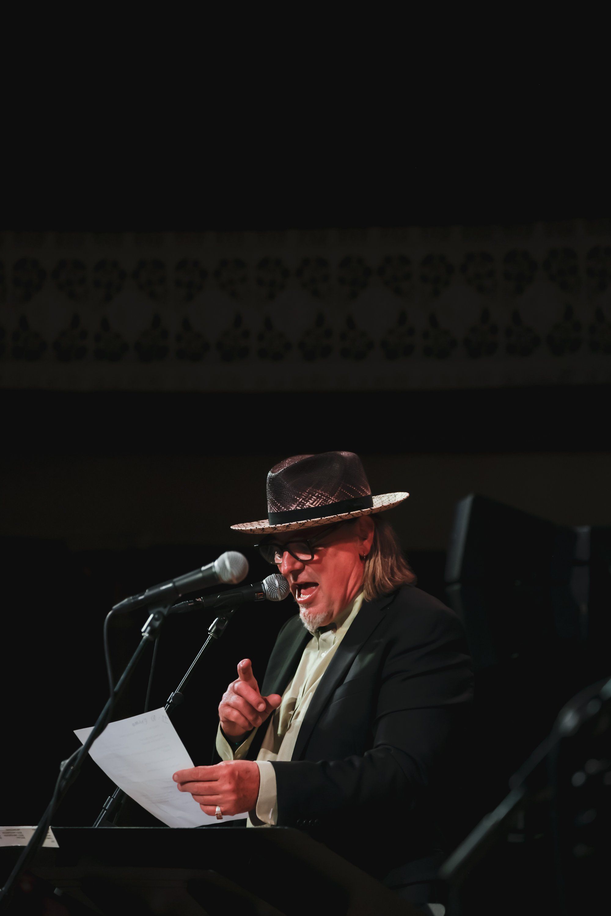 Man in cowboy hat and dark jacket speaks passionately at microphone, holding papers, against dark stage backdrop