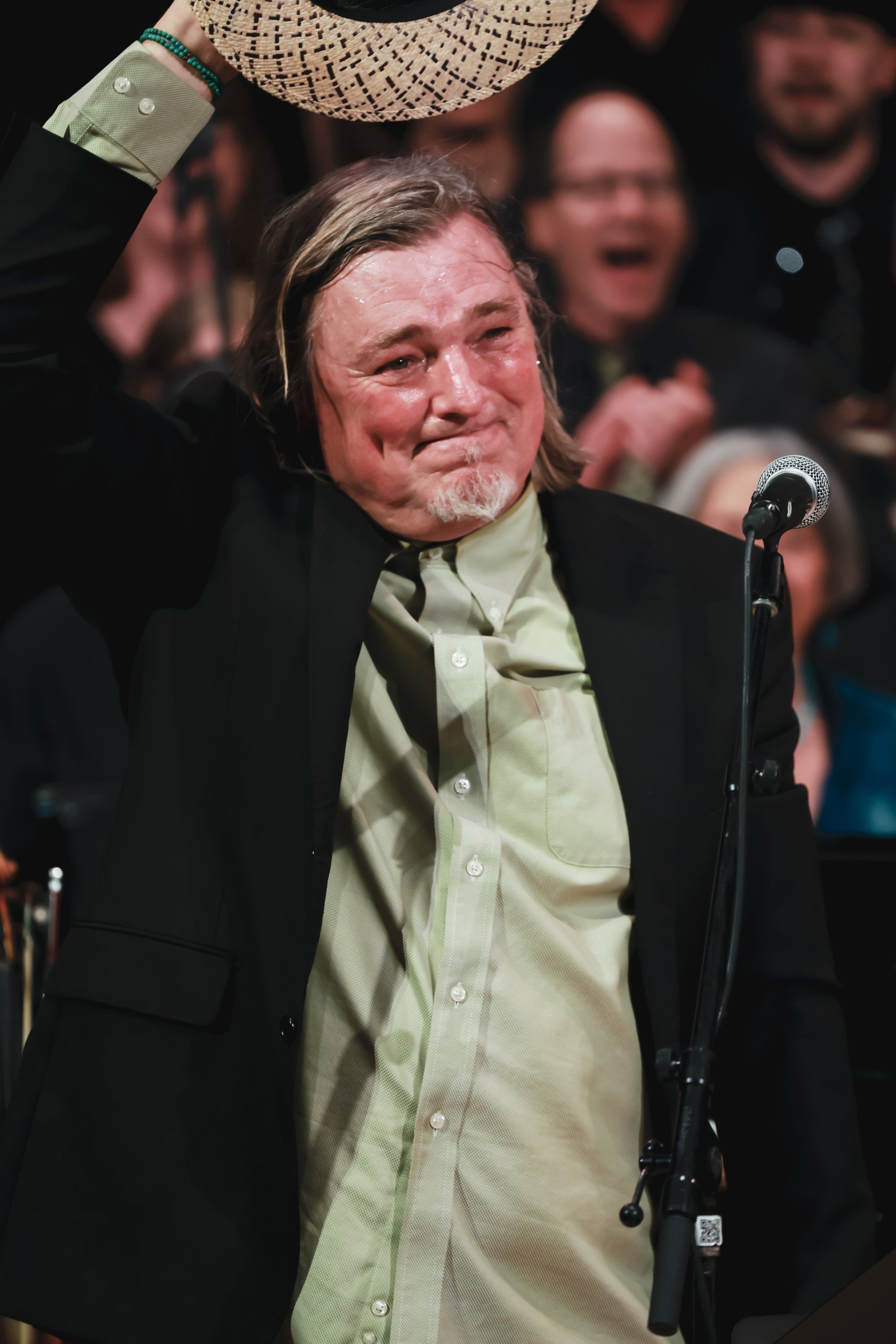 Man in cream shirt and black blazer holding award aloft at microphone, smiling amid applauding audience