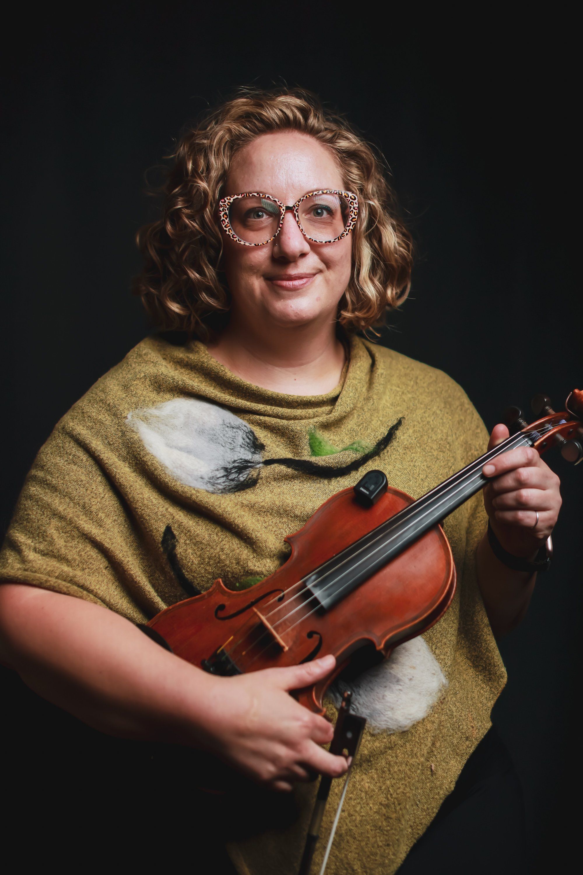 Young violinist in golden-yellow shirt and glasses holds instrument against dark background, warm studio lighting
