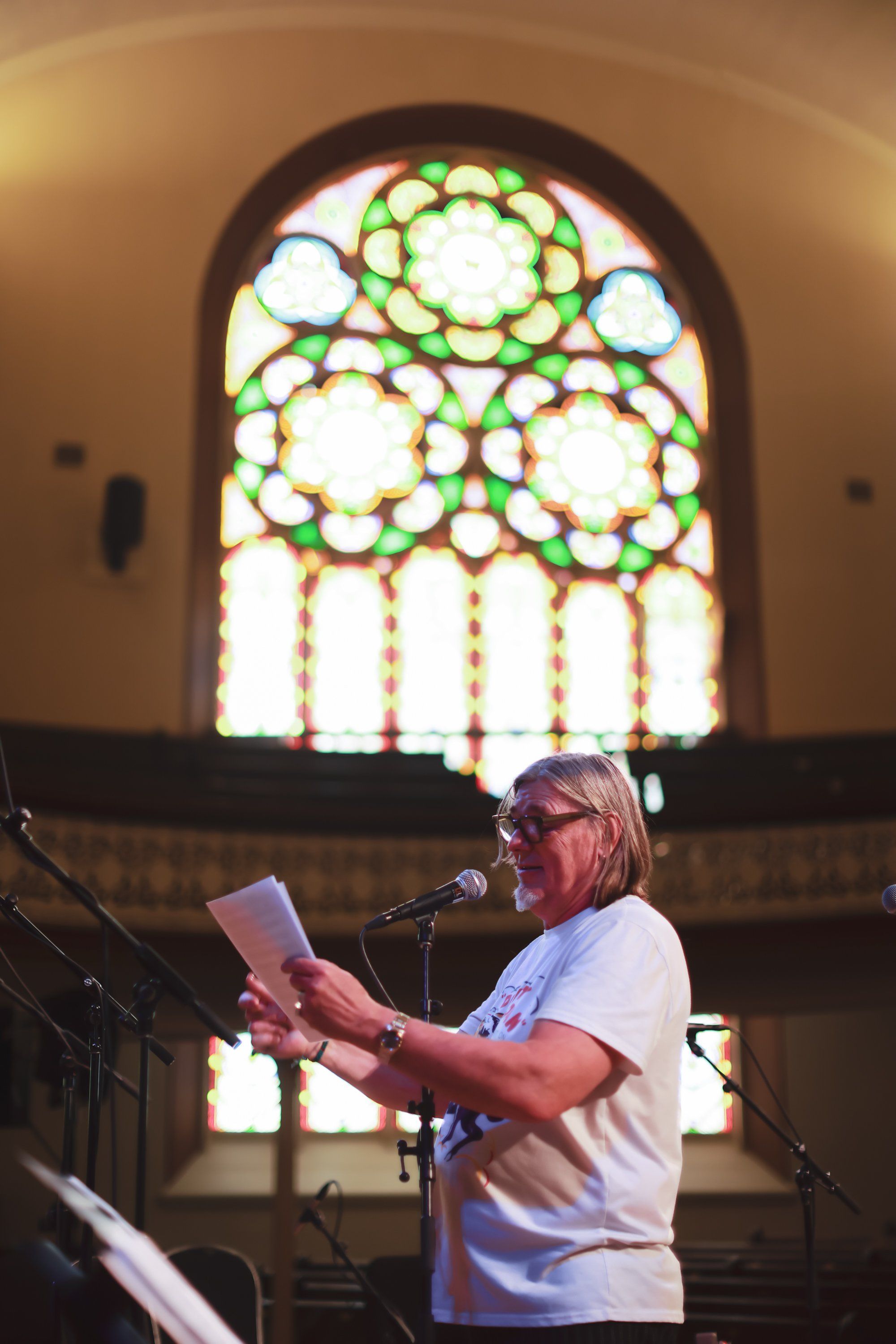 Musician at microphone in white shirt reading from paper, backlit by stained glass window with green and white geometric patterns in church interior