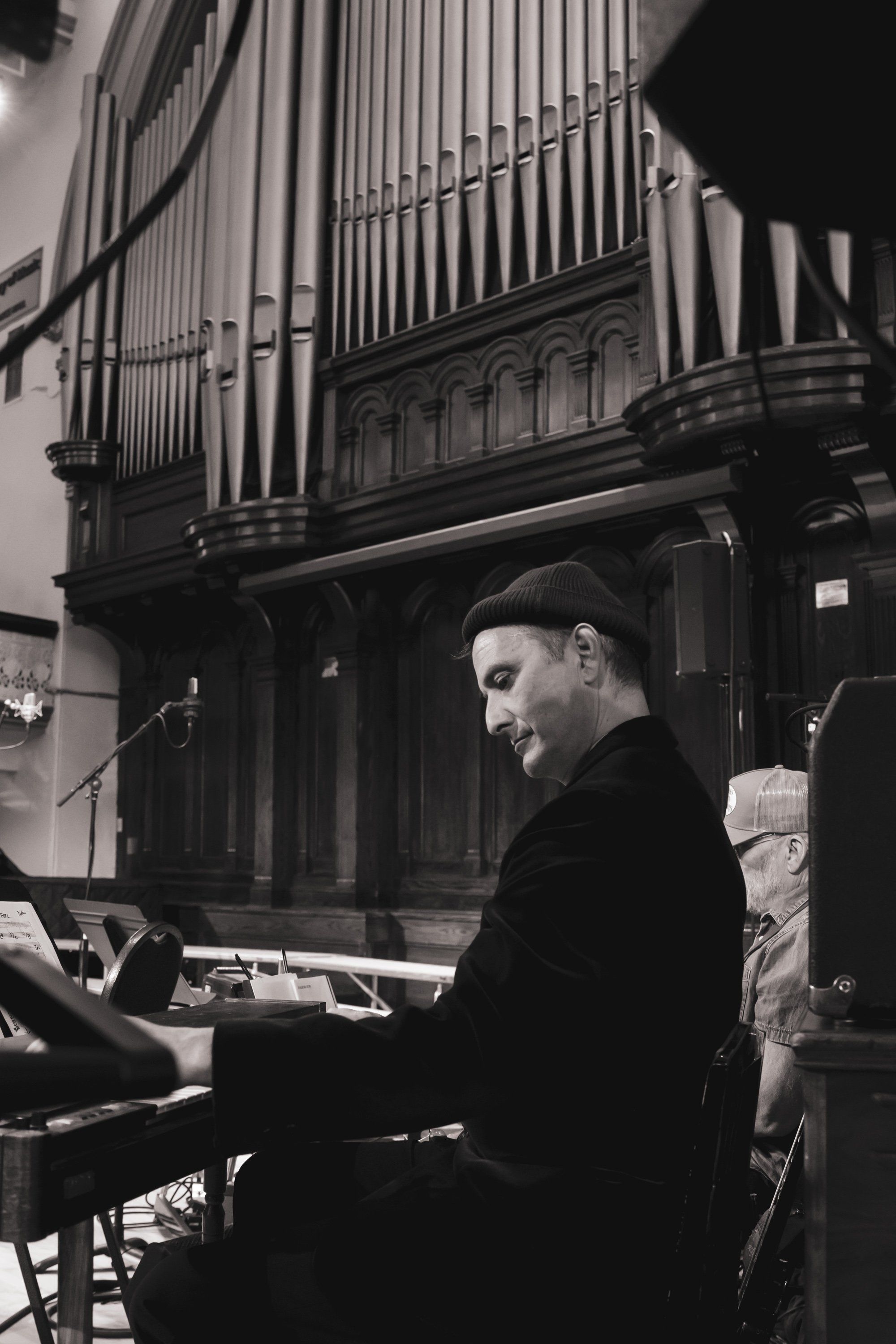 Musician seated at organ console in dramatic black and white, ornate pipe organ visible behind in concert hall setting