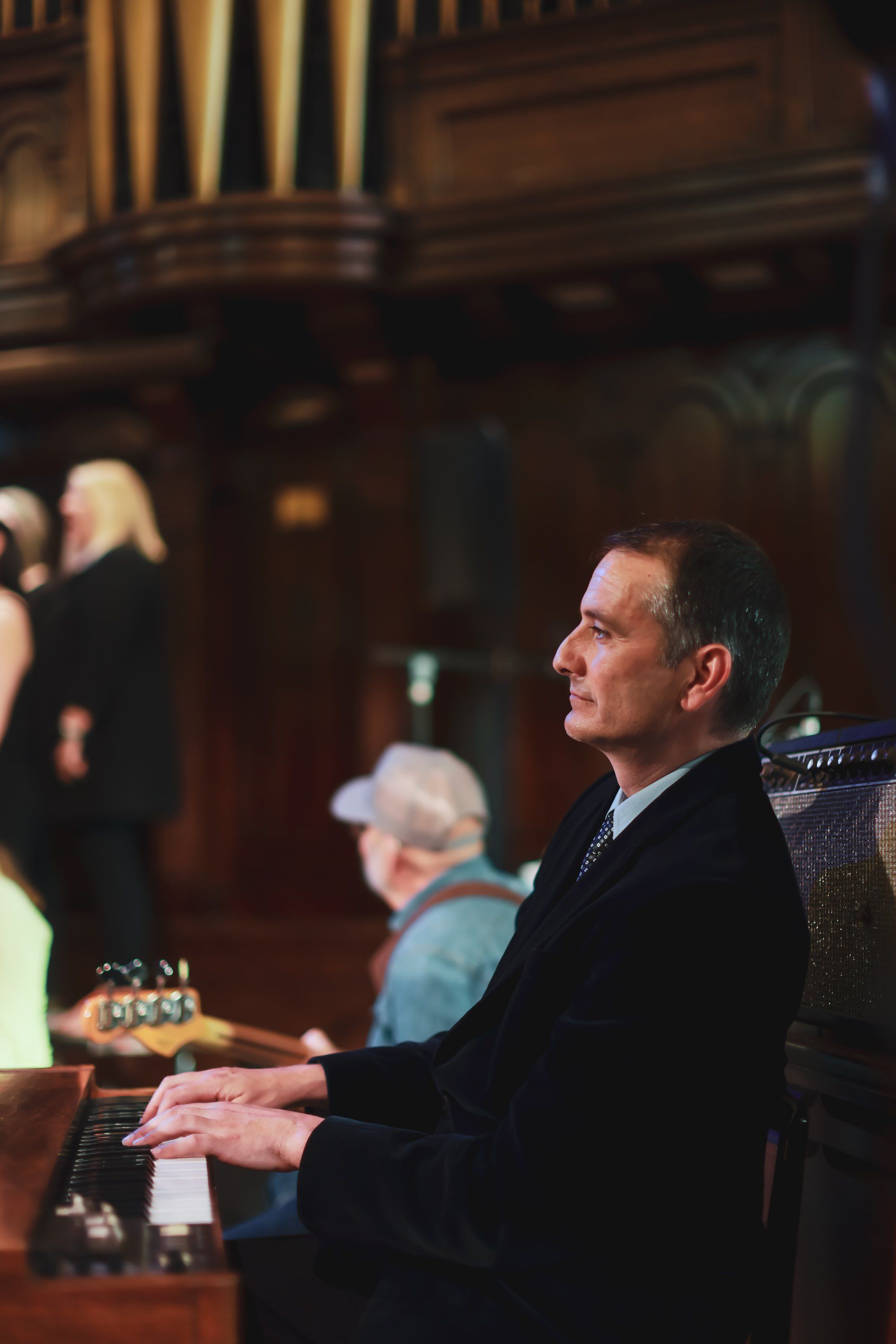 Man in dark suit seated at piano in ornate wooden interior, warm stage lighting, contemplative expression