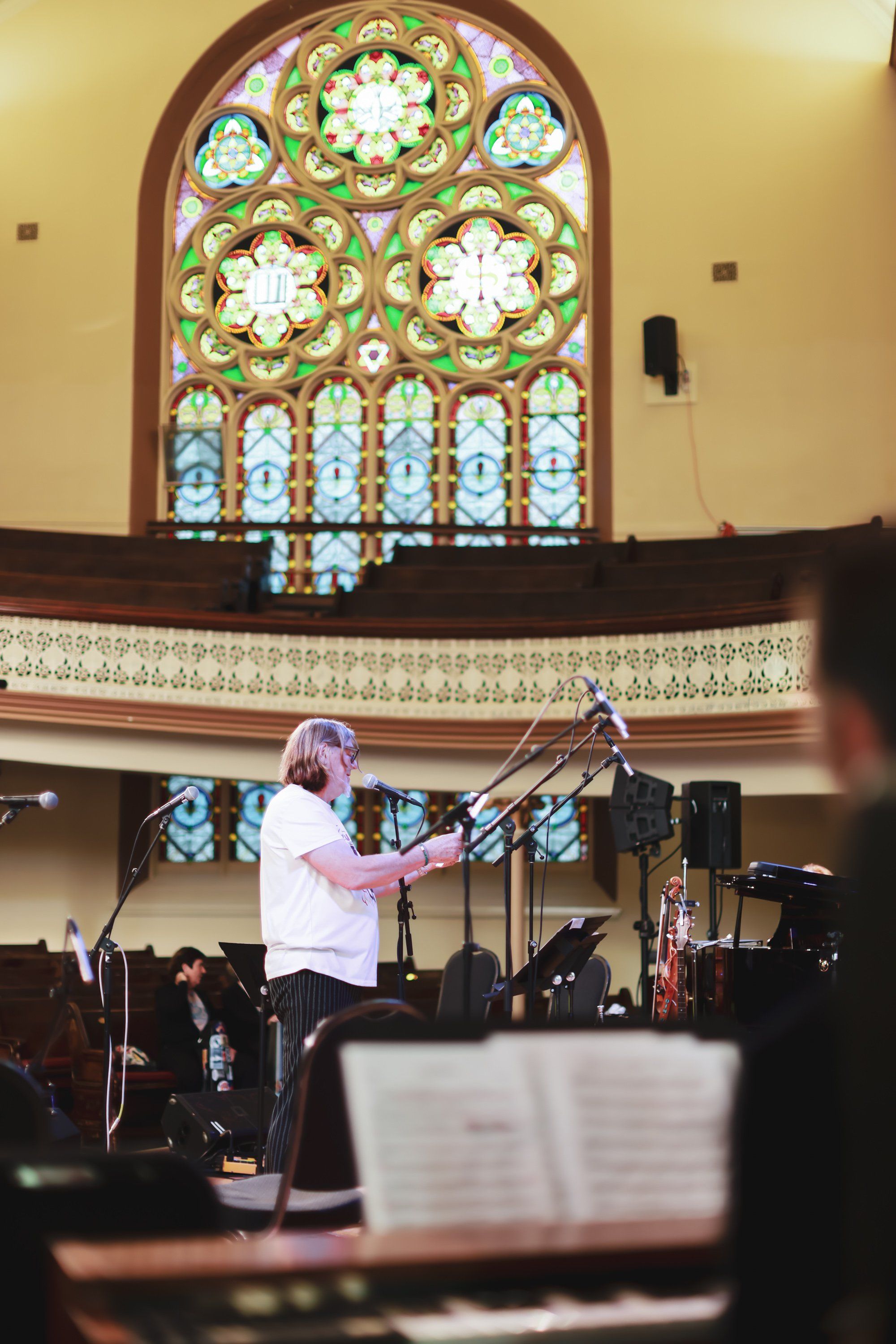 Conductor in pink shirt leads orchestra beneath ornate stained-glass window in warm-lit church sanctuary