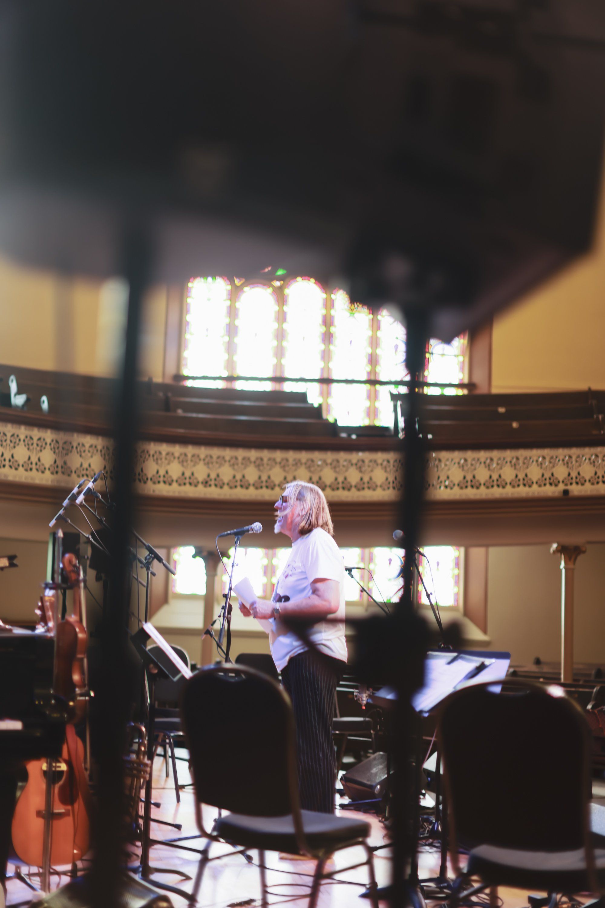 Musician in light purple shirt performs in ornate concert hall with dramatic backlighting through tall windows, silhouetted audience in foreground