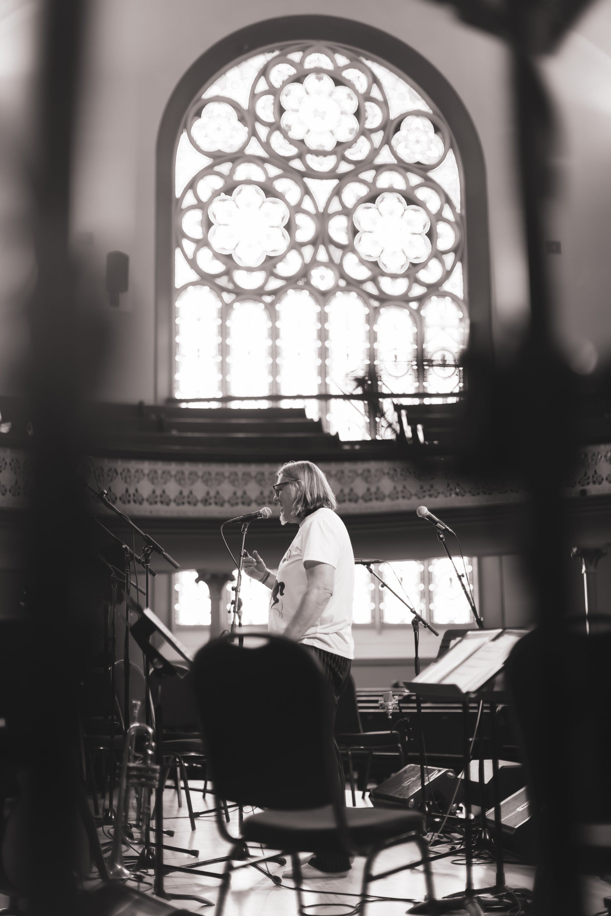 Musician with long hair seated at keyboard, backlit by ornate arched window with geometric tracery in concert hall