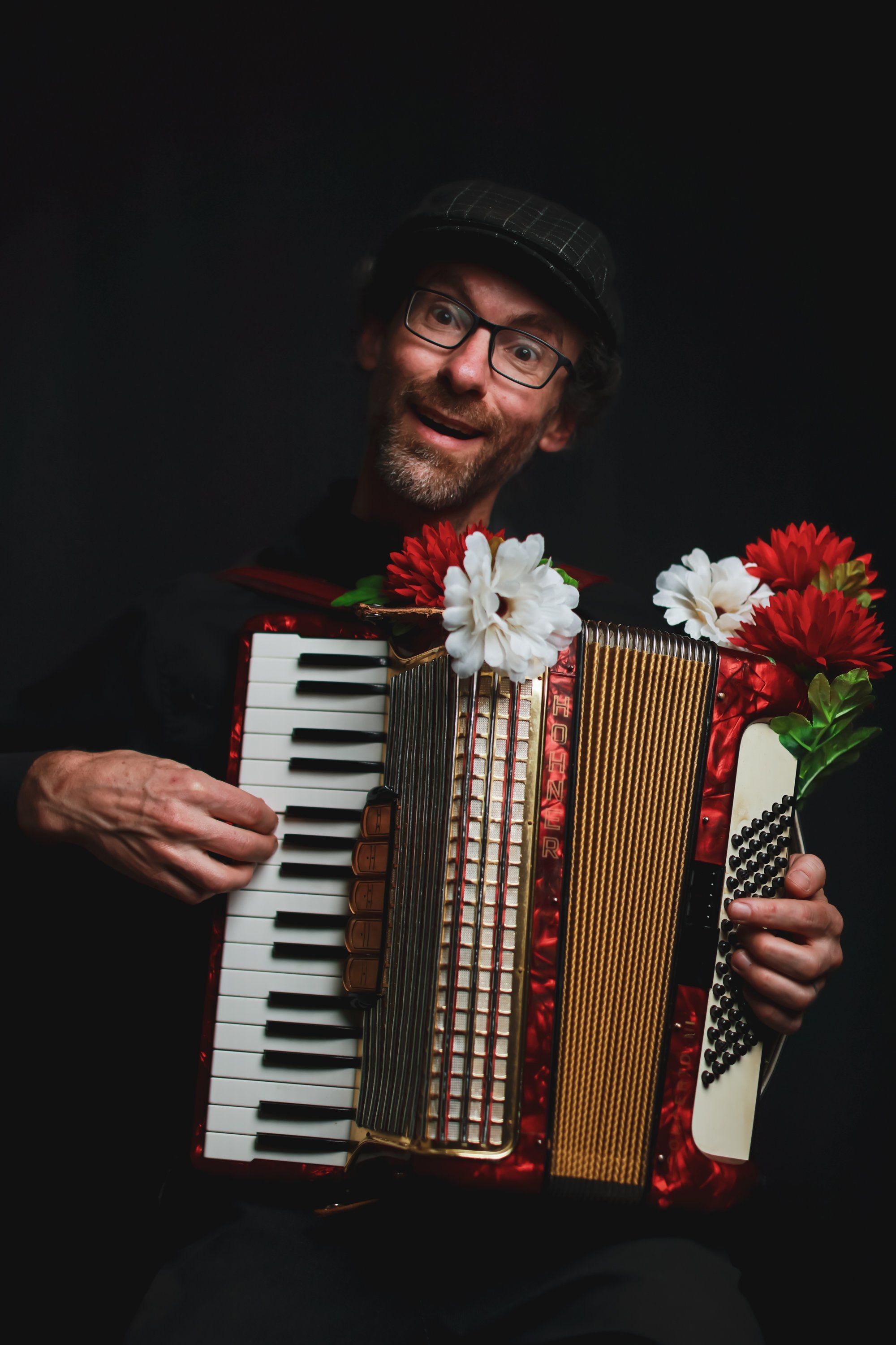 Smiling musician with glasses and beanie holds a red accordion adorned with red and white flowers against a black background