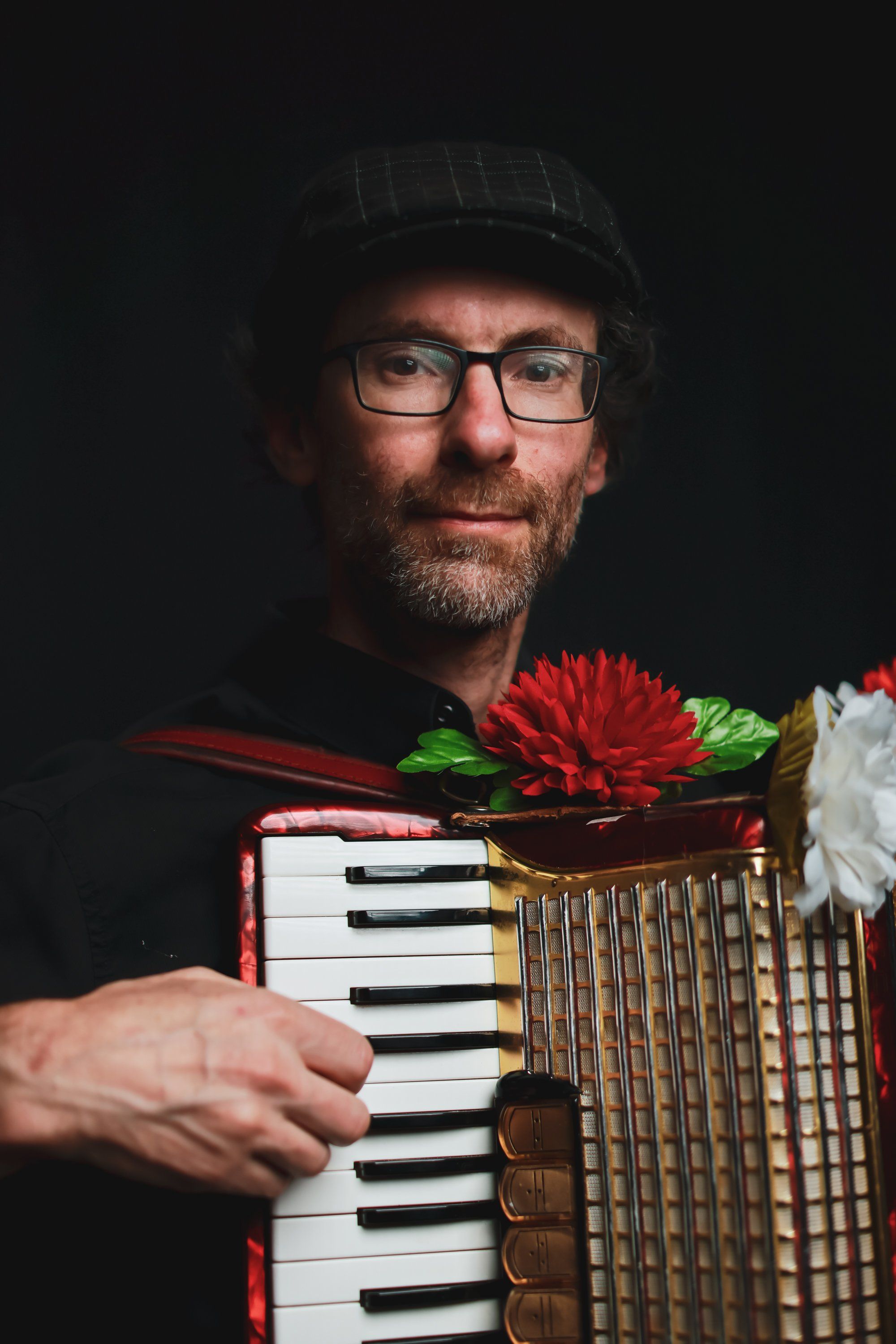 Bearded musician in glasses and beanie holds a red accordion decorated with flowers against a dark background