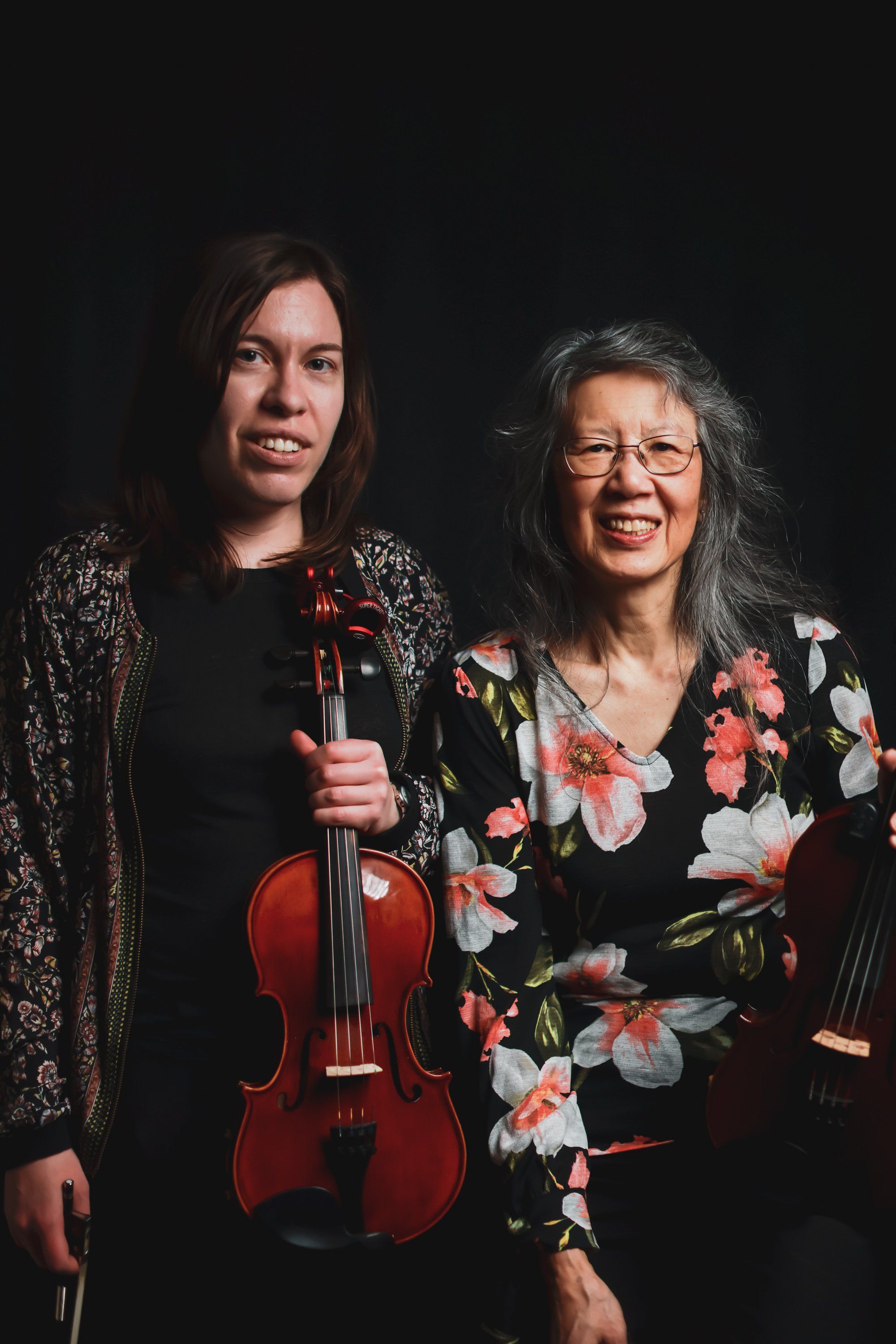 Two musicians against a black backdrop: a younger woman in a sparkly jacket holding a violin, and an older woman in a floral black dress, both smiling warmly…