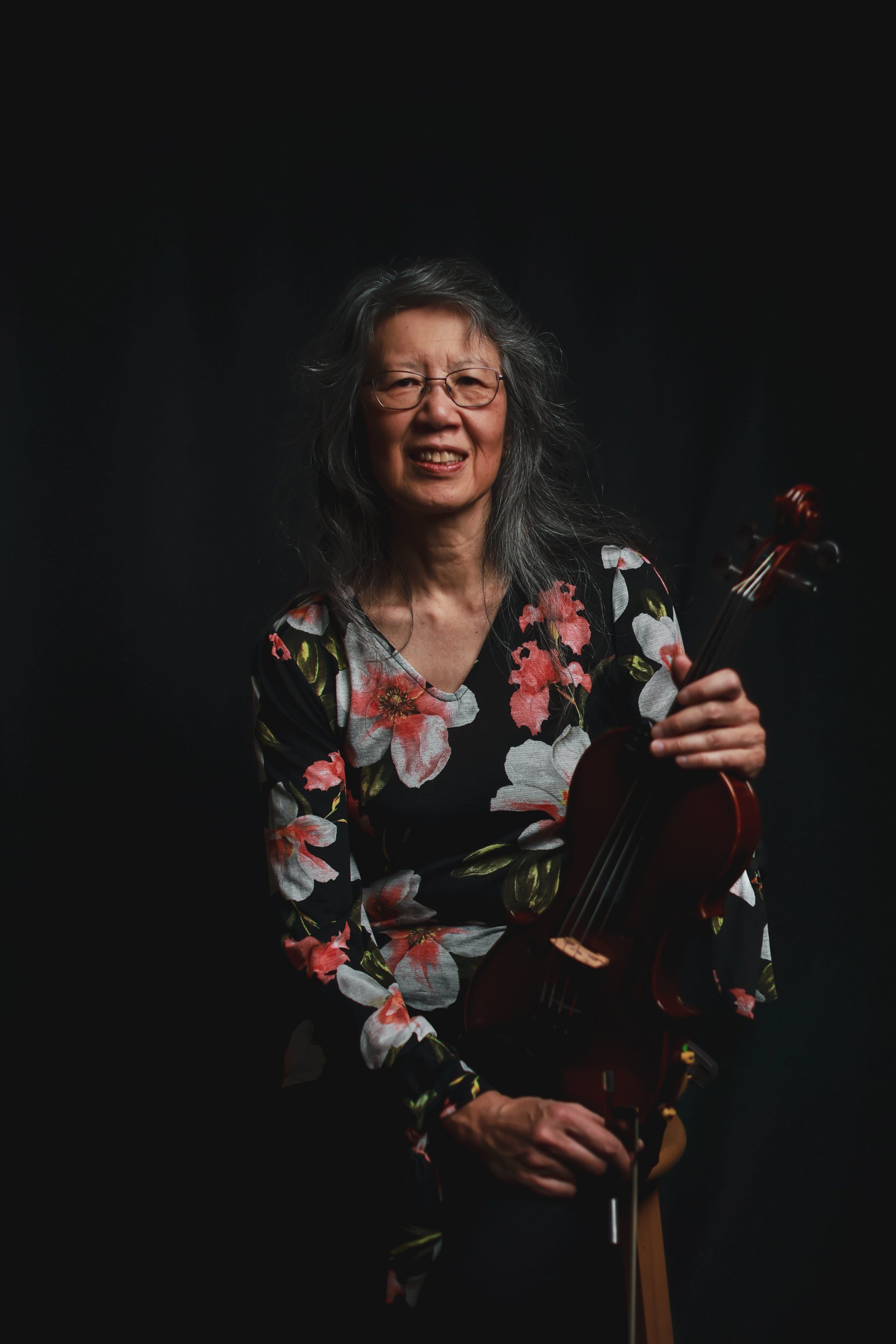Smiling musician with long dark hair and glasses holds a violin against a black backdrop, wearing a vibrant floral print jacket