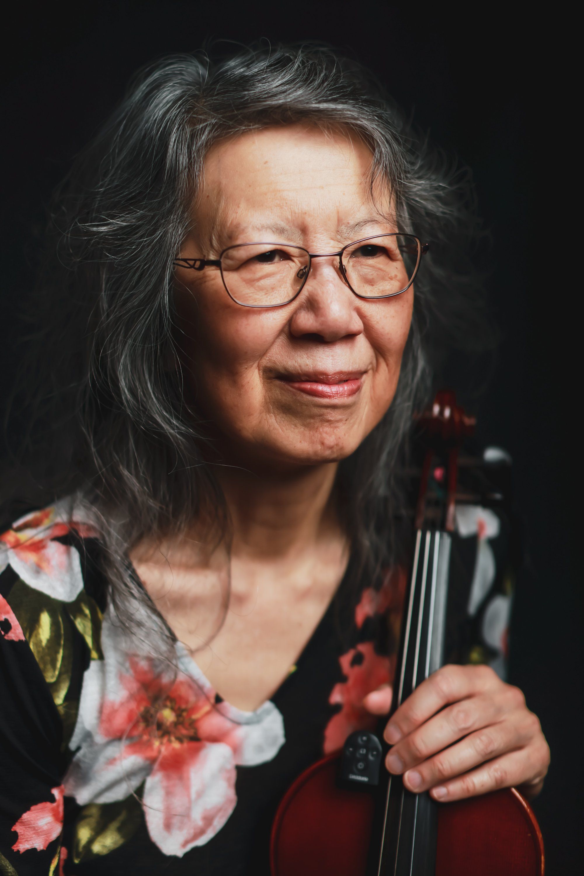 Violinist with long gray hair and glasses holds her instrument against a dark background, wearing a floral patterned dress and smiling warmly at the camera