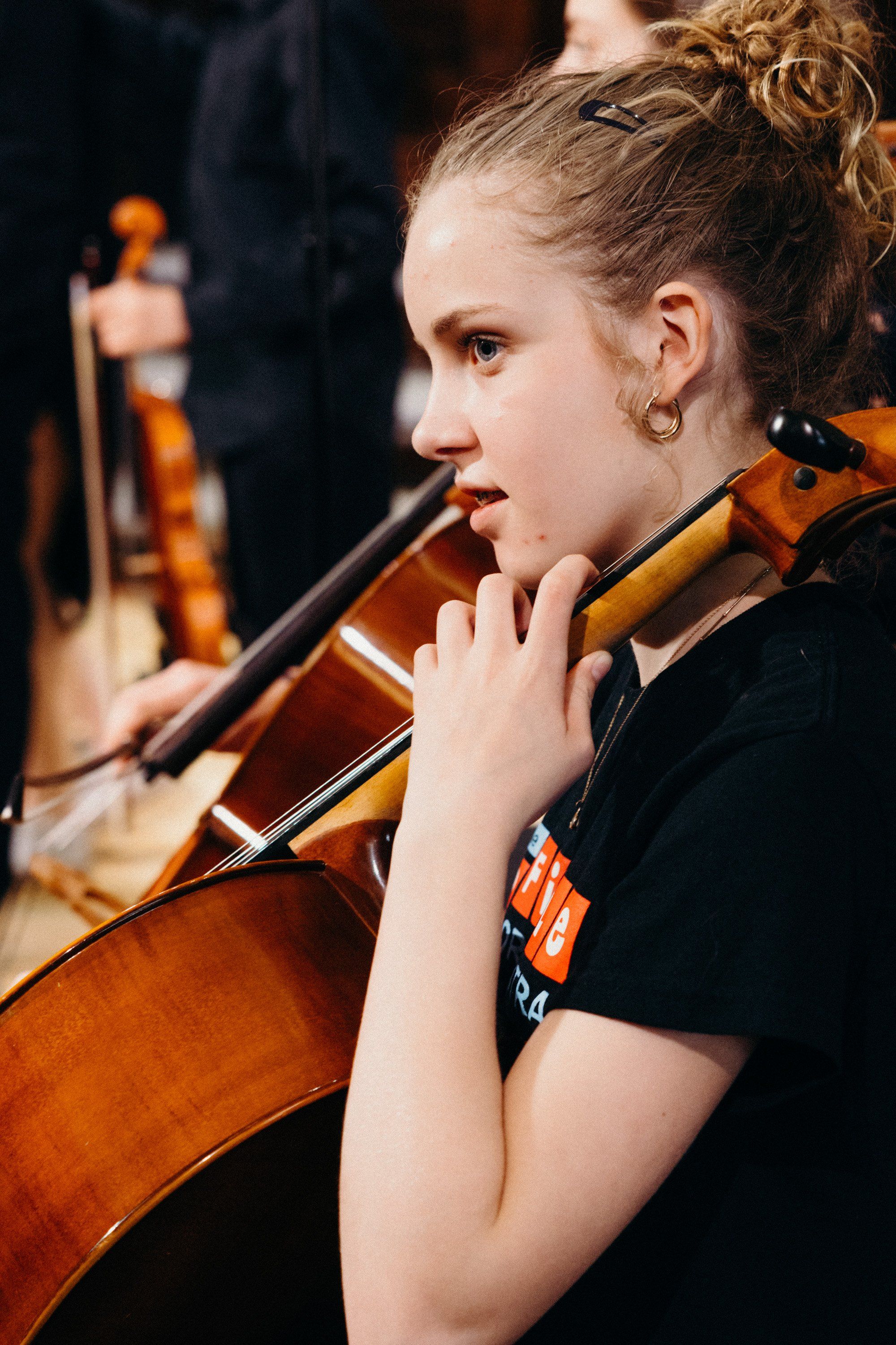 Young cellist in black performance attire concentrating intently while playing, warm stage lighting illuminating her focused expression and wooden instrument