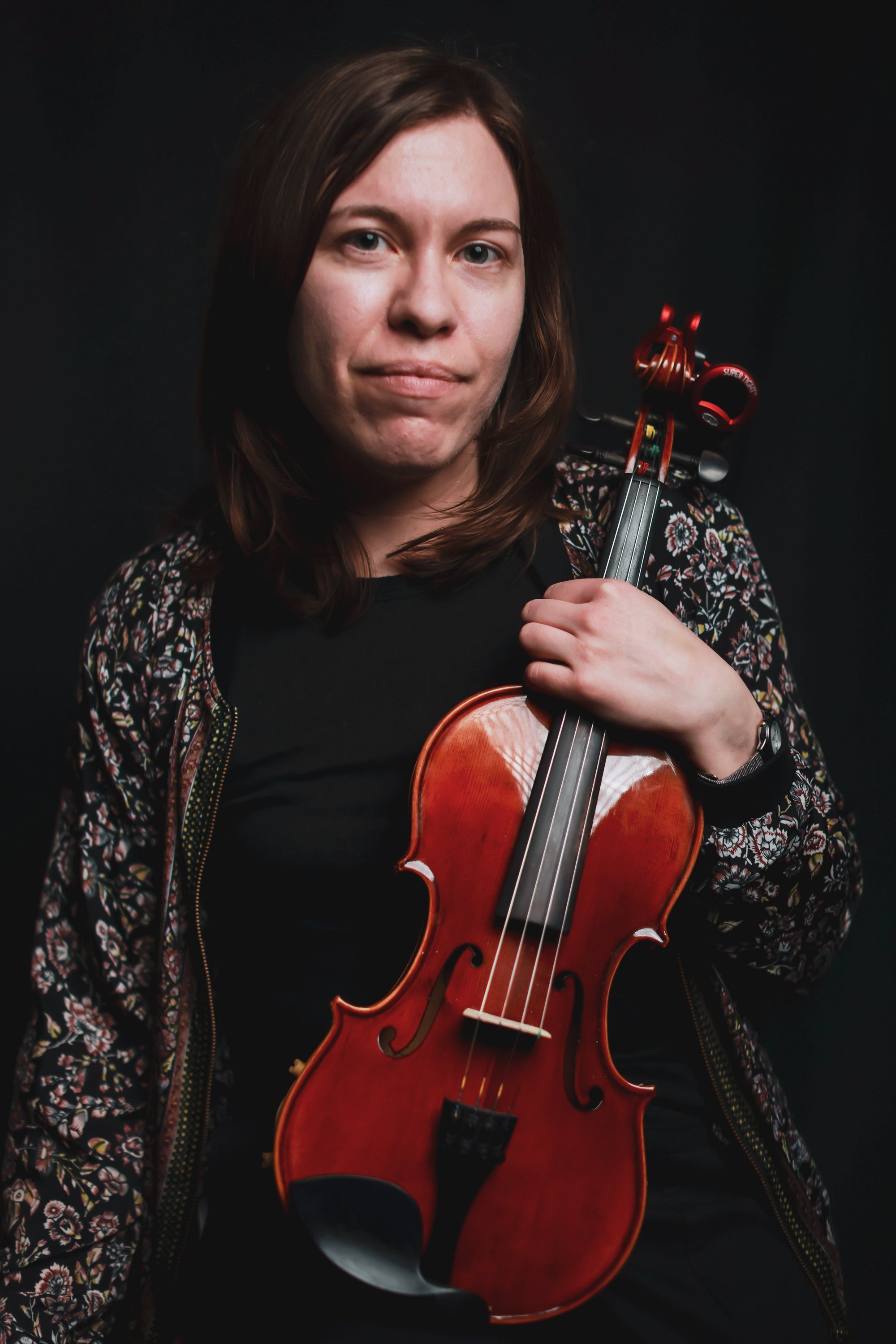 Woman holding a red violin against a dark background, wearing a patterned cardigan over black, with soft directional lighting