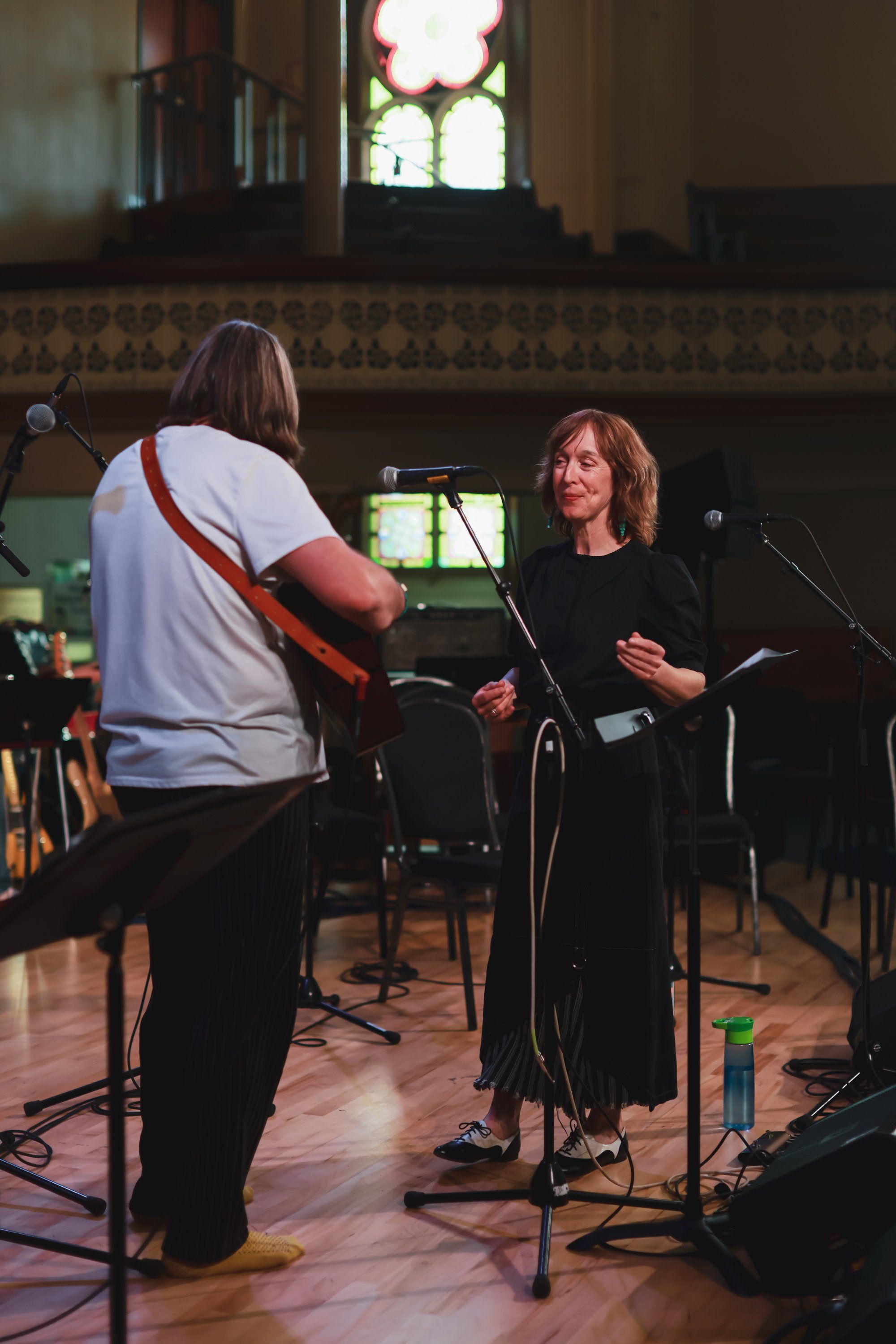 Musician with guitar performing indoors while woman in black speaks at microphone in a wooden-beamed hall with stained glass window
