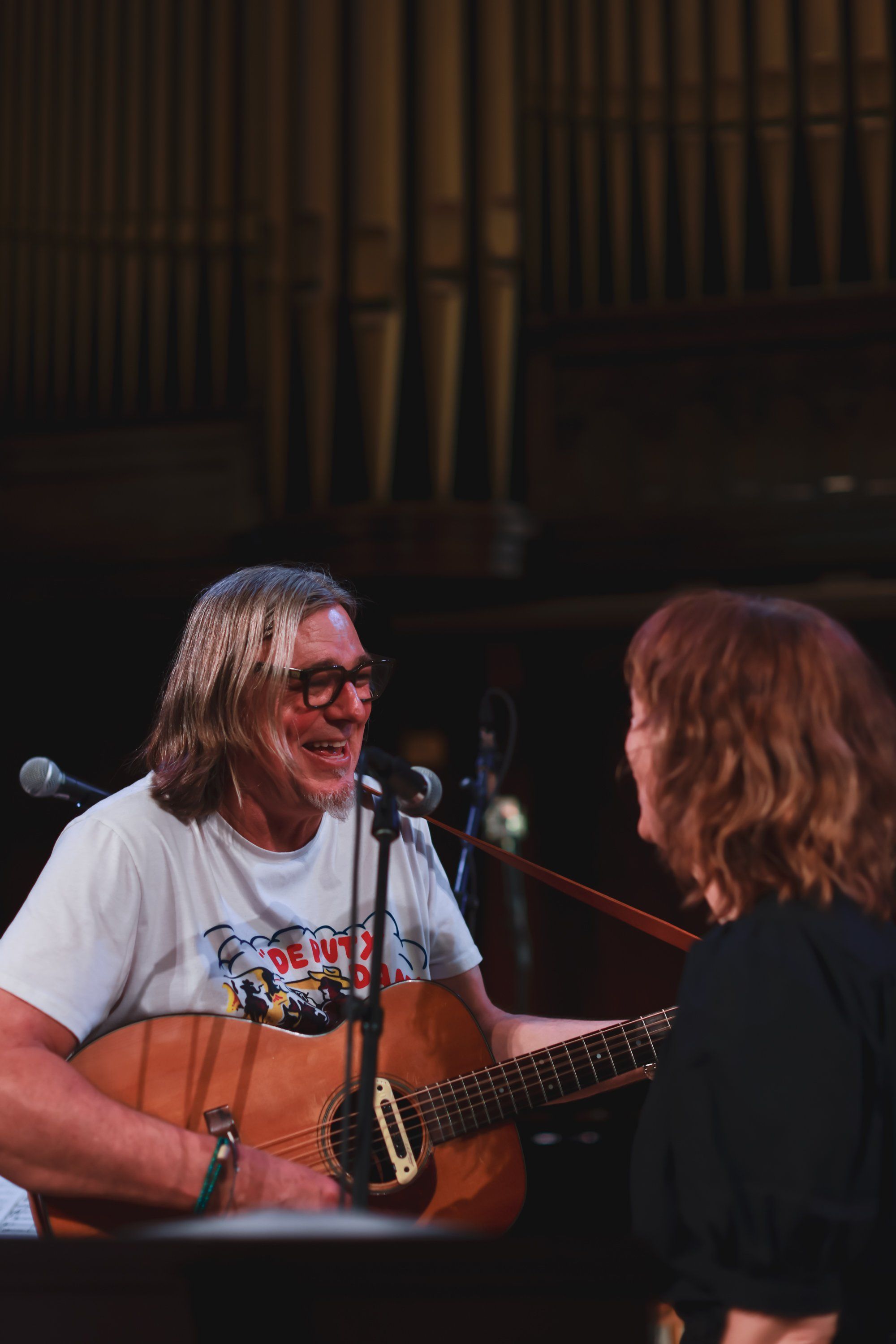 Musician with long hair and glasses performs with acoustic guitar at a concert, smiling toward a female performer beside him on stage