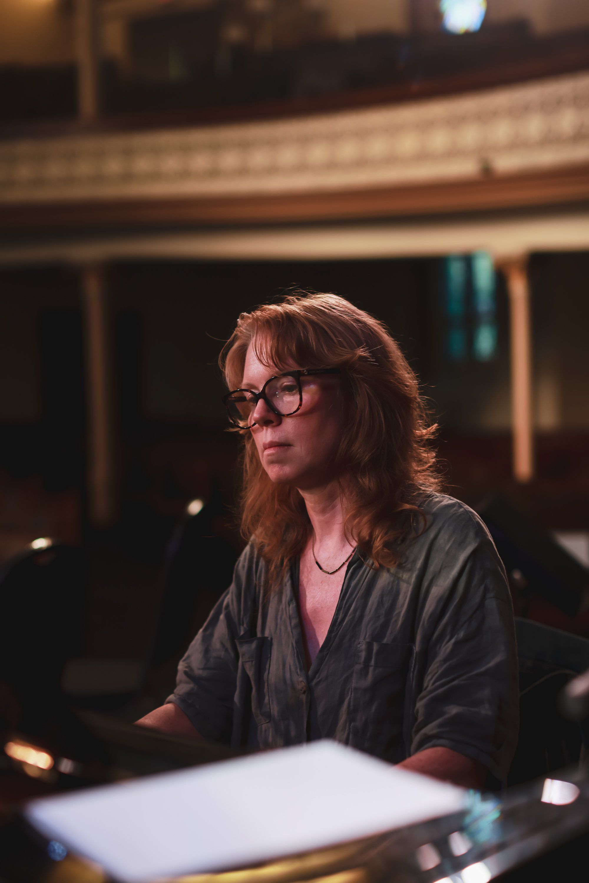Woman with glasses and reddish-brown hair sits at a piano in warm stage lighting, gazing thoughtfully to the side