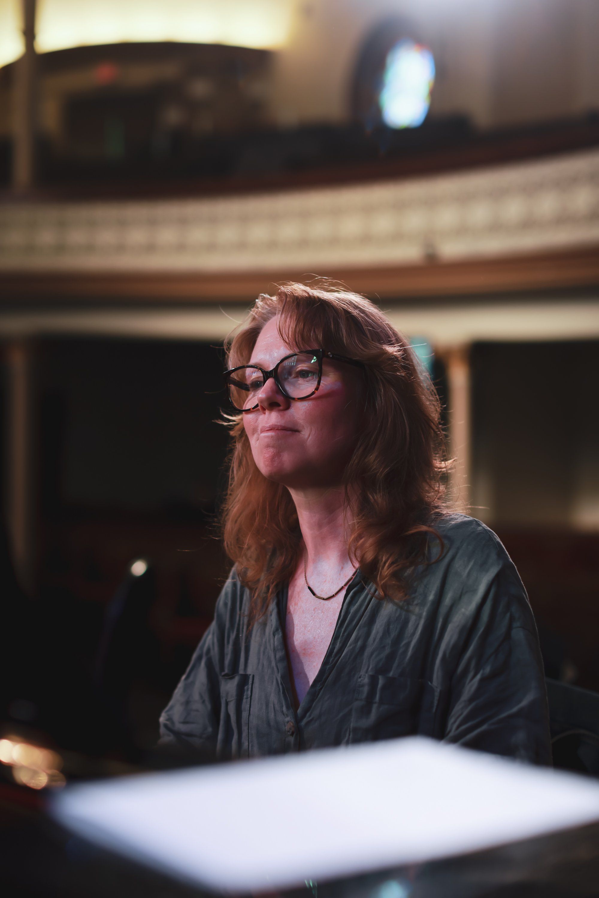 Woman with glasses seated at a concert venue, lit by warm stage lighting, gazing upward with contemplative expression