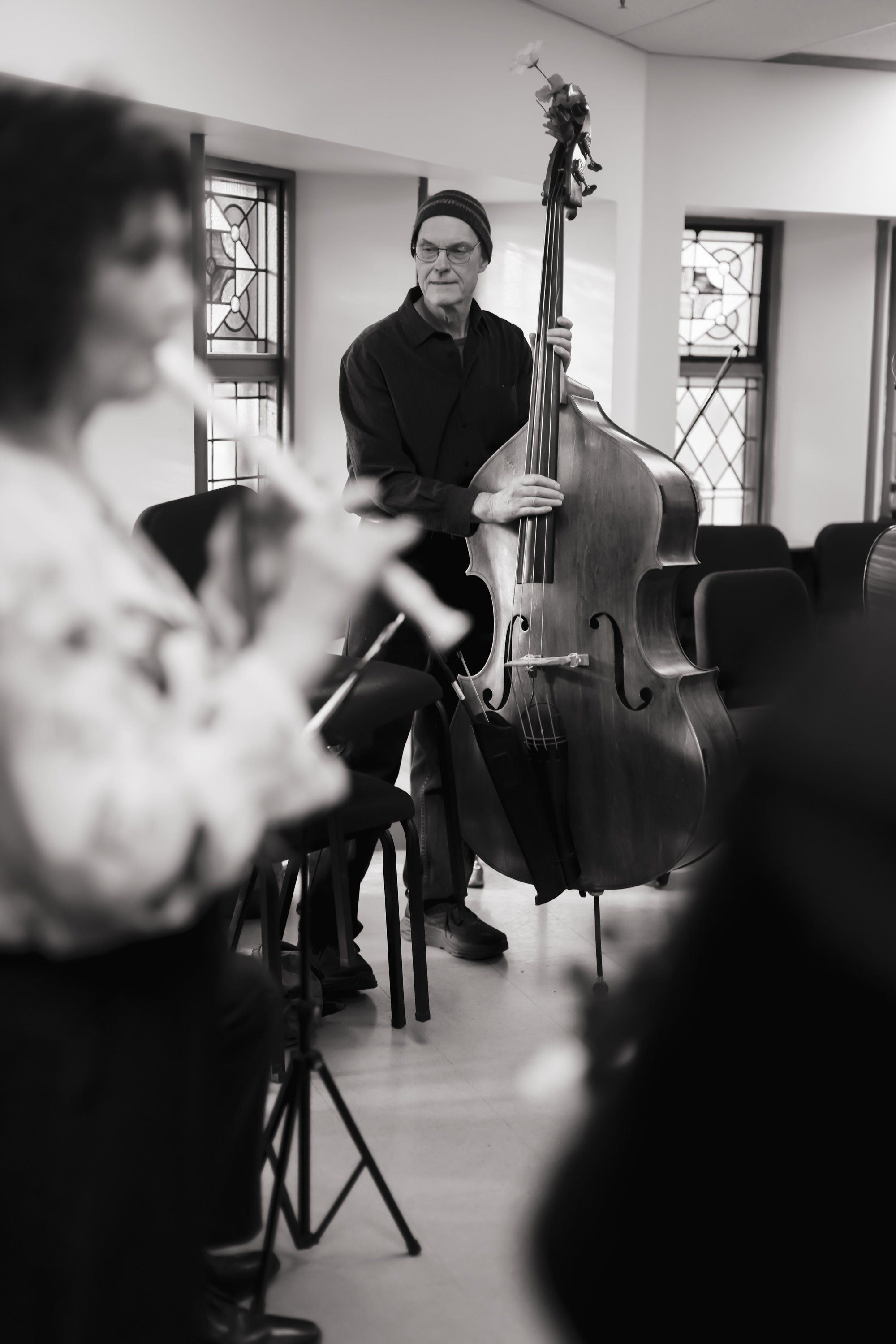 Musician with upright bass in concert hall, focused expression, soft natural light through windows, other performers blurred in foreground