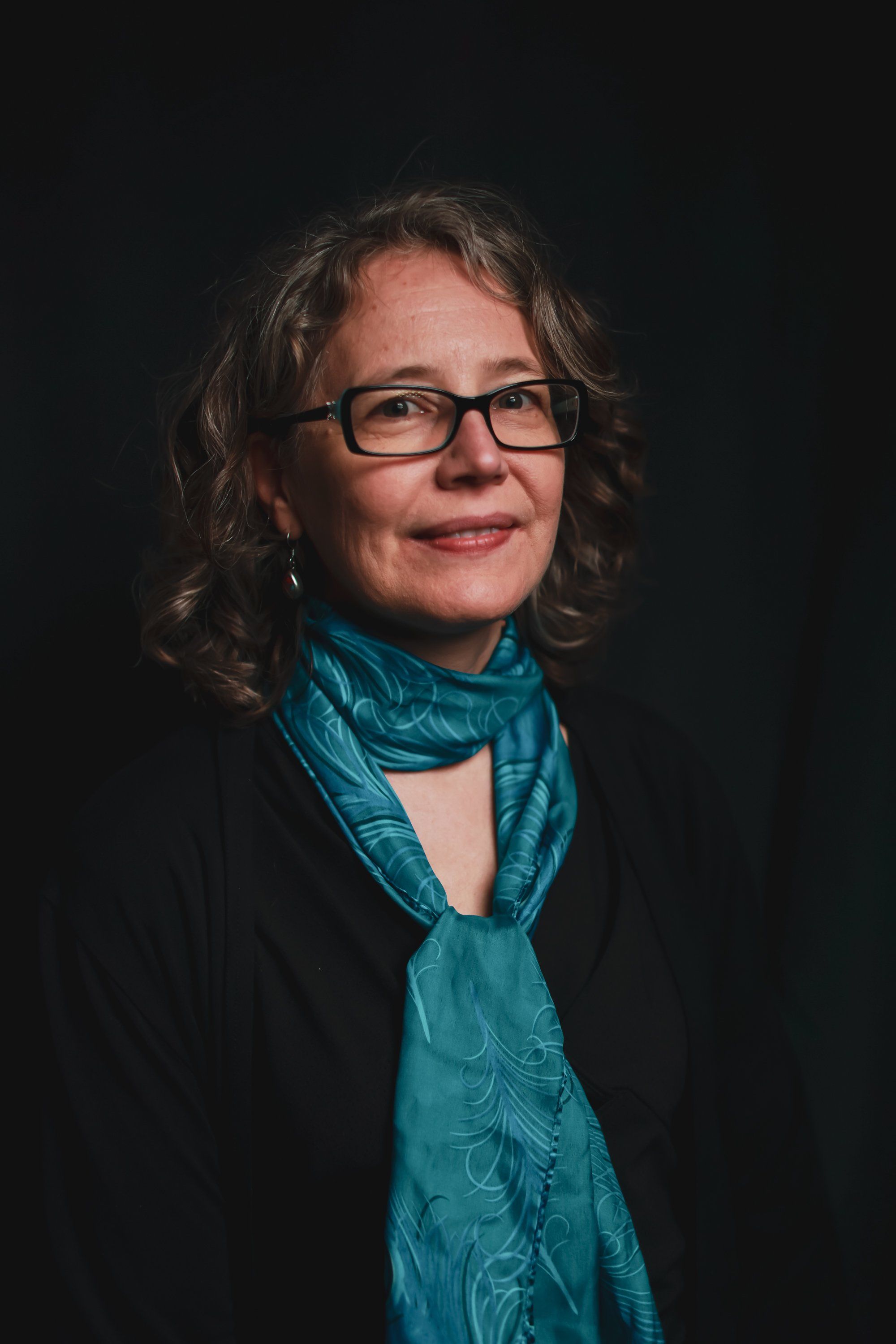 Woman with shoulder-length wavy hair and glasses wearing a turquoise scarf and black jacket, photographed against a dark background