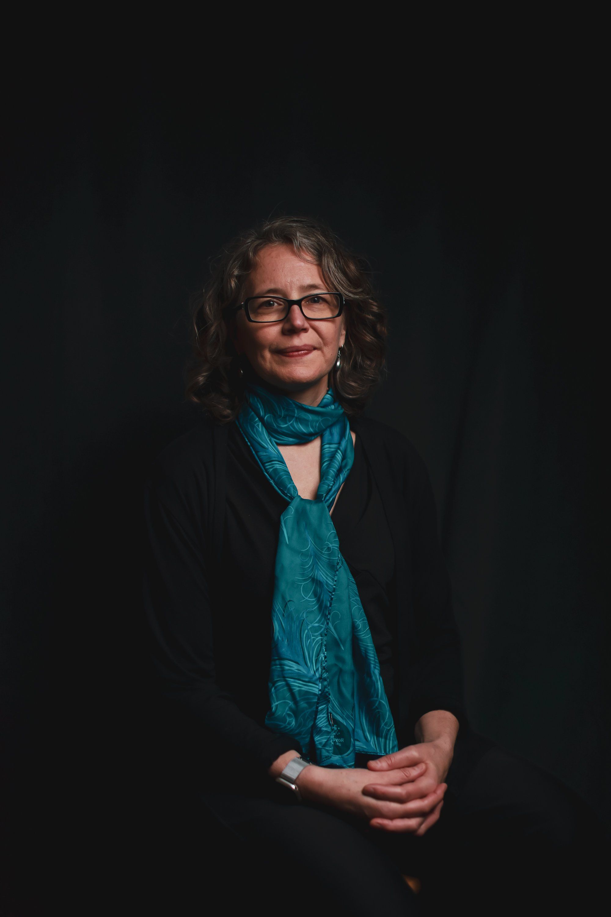 Woman with glasses and shoulder-length hair wearing a turquoise scarf against a dark background, hands clasped in front
