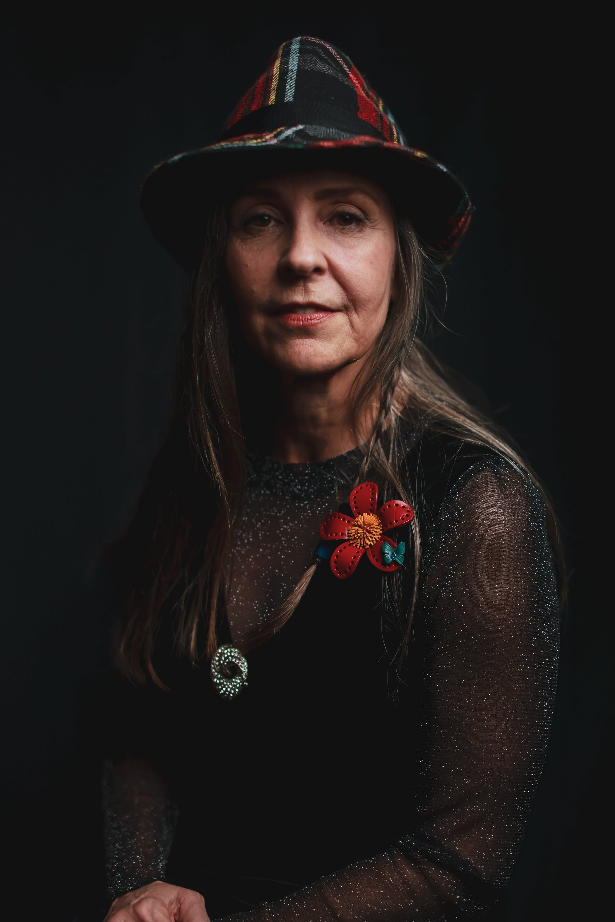 Woman with long dark hair wearing a red plaid hat and black mesh top with red flower brooch, photographed against dark background