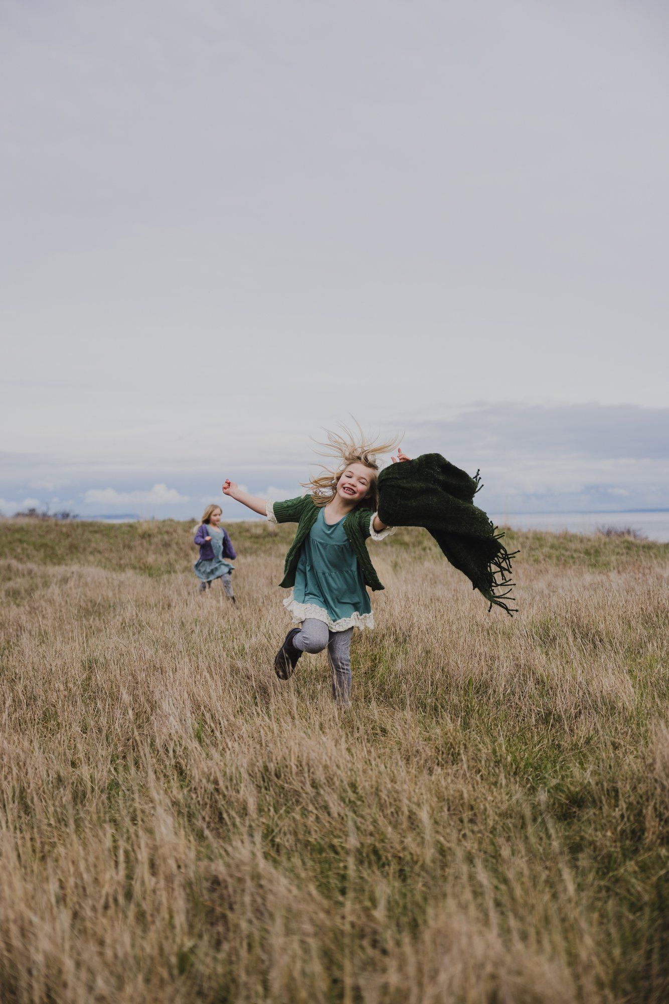 Woman in green dress running through golden grassland with child, mountains hazy in distant background