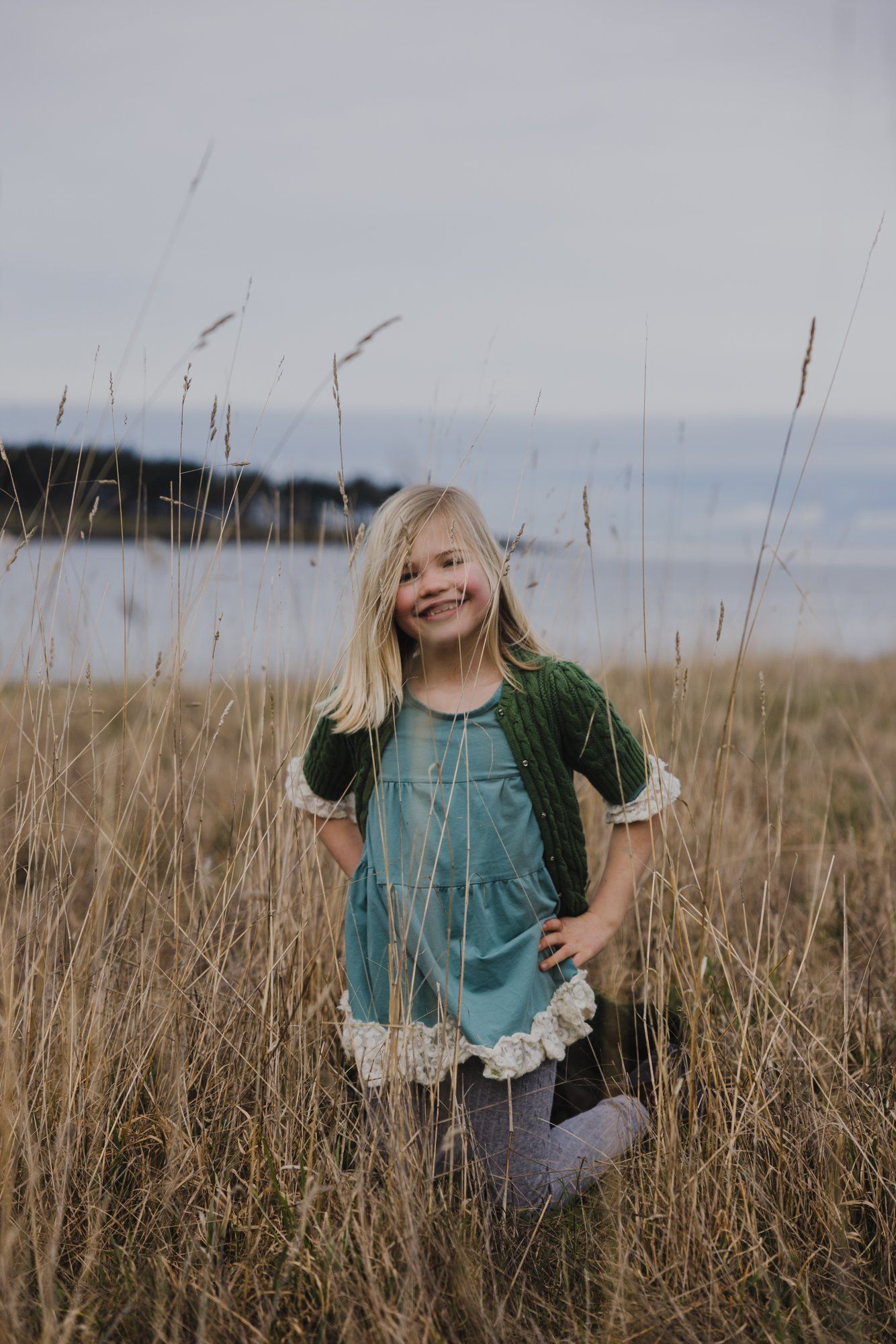 Young girl with blonde hair smiling in tall dried grasses with water and shoreline visible behind her
