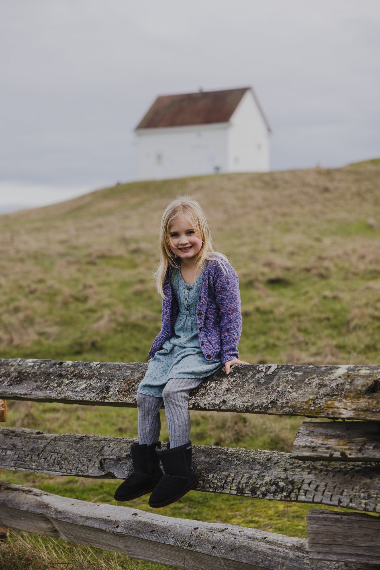 Young girl sitting on weathered fence rails with rolling grassland and small house on hillside behind her