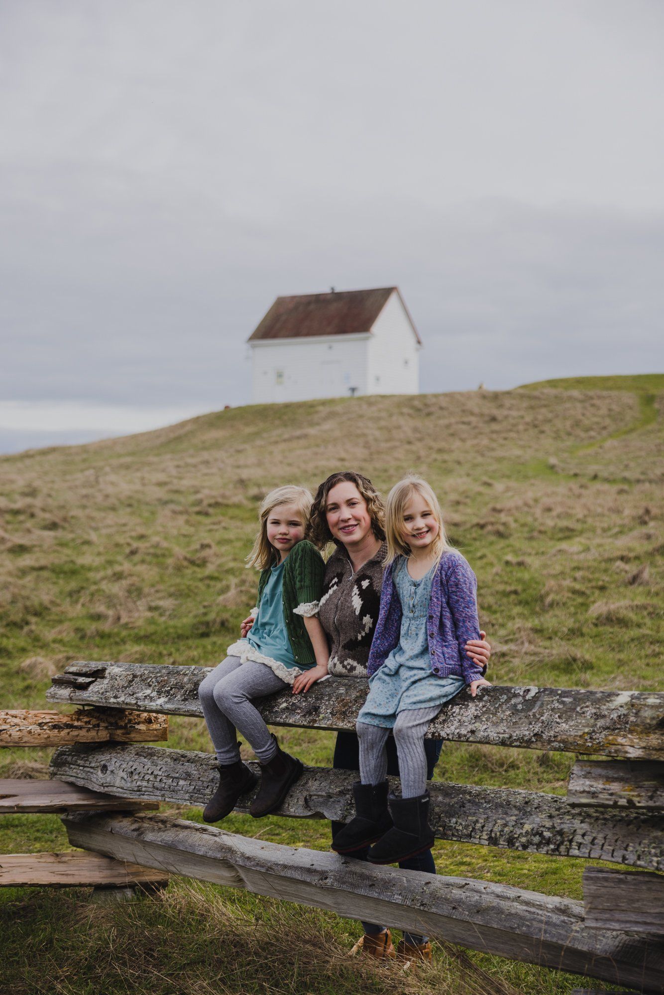 Three girls sit on weathered driftwood logs on a grassy hillside with a white farmhouse and overcast sky behind them